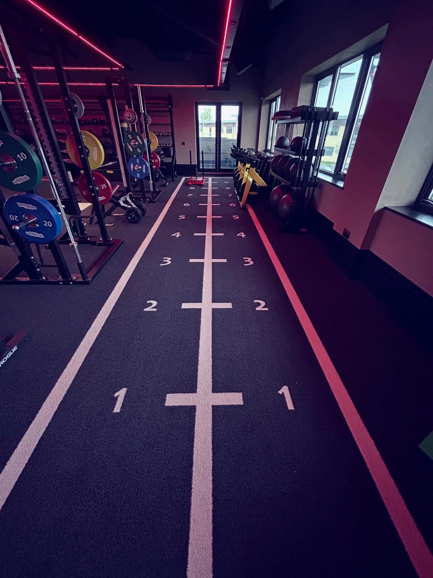 Indoor gym with weightlifting equipment and a marked running track on the floor, illuminated with pink LED strip lighting.