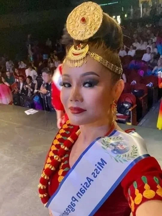 A woman dressed in traditional attire taking a selfie on stage during a cultural event, with an audience seated in the background.