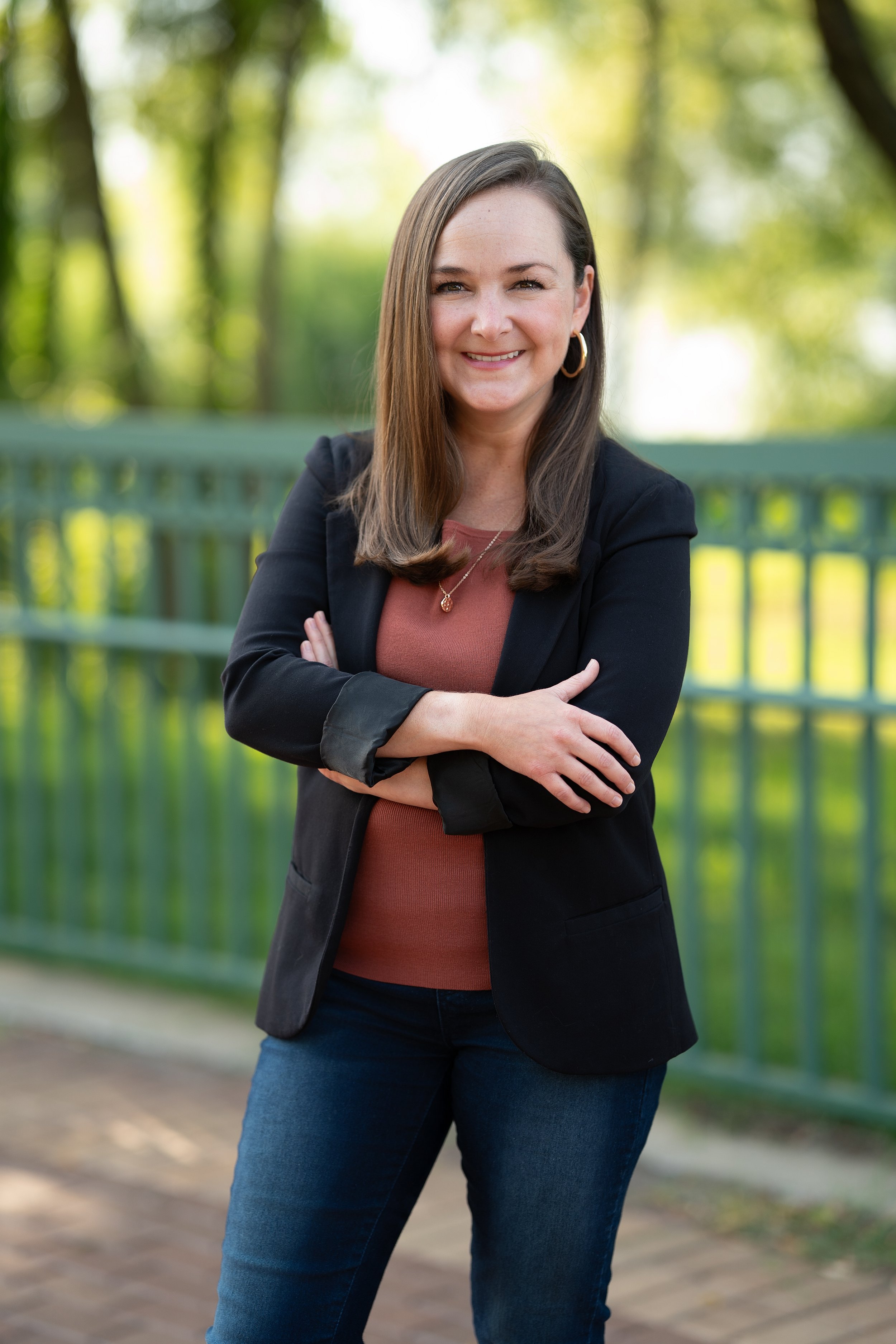 A smiling woman with brown hair, wearing a black shirt, teal blazer, and blue jeans, stands outdoors at an area with string lights and benches.