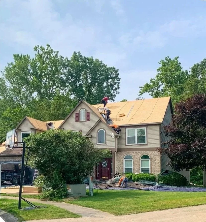 Construction workers installing a new roof on a two-story house with beige vinyl siding and brick facade, in a suburban neighborhood with green trees and a basketball hoop.