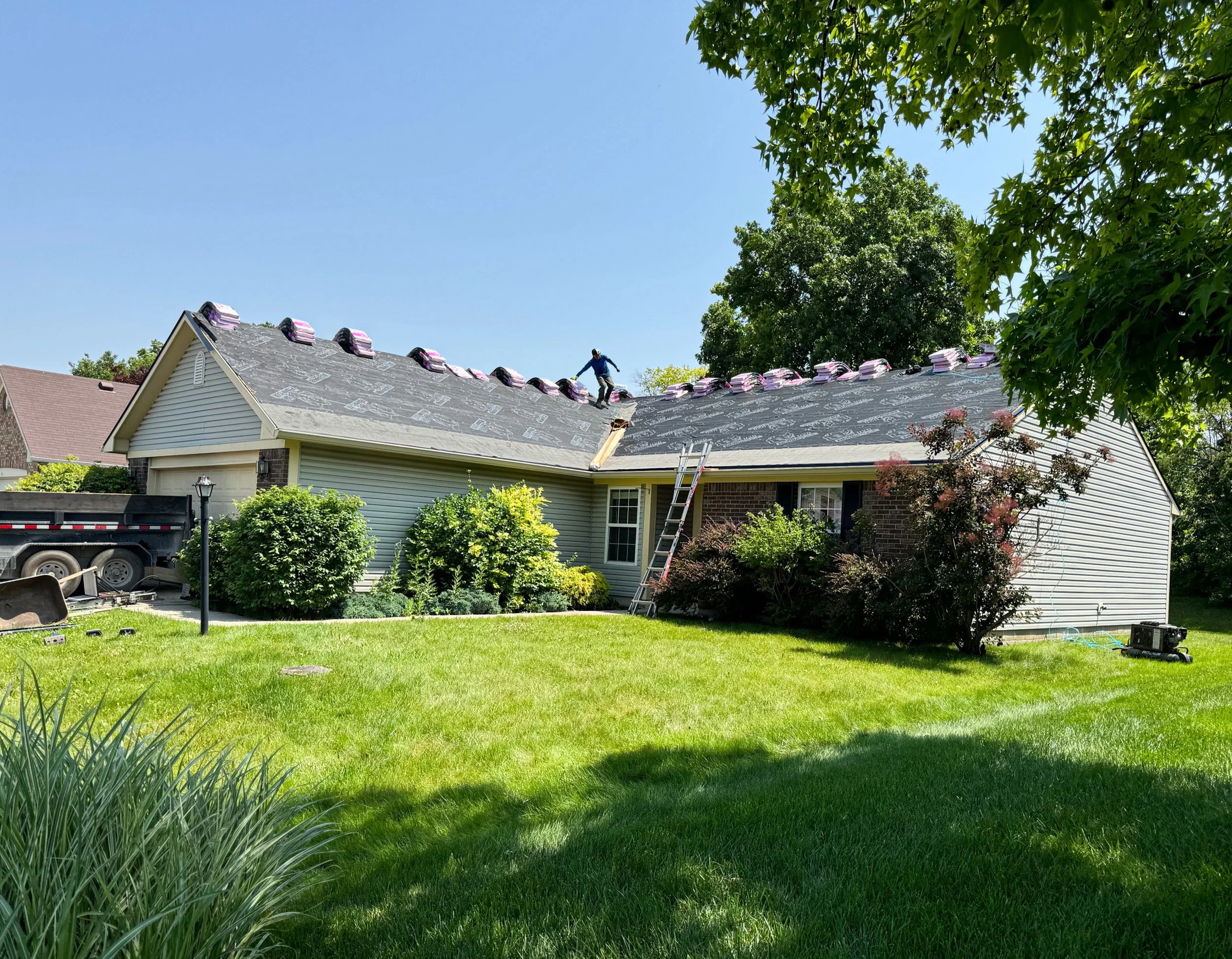 A house undergoing roof replacement with workers installing new roofing materials. The house is surrounded by a well-maintained yard with trees and bushes, and a ladder leans against the house for access.