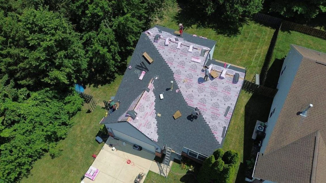 Aerial view of a house roof being renovated, with several workers installing new shingles and insulation, surrounded by green trees and neighboring houses.