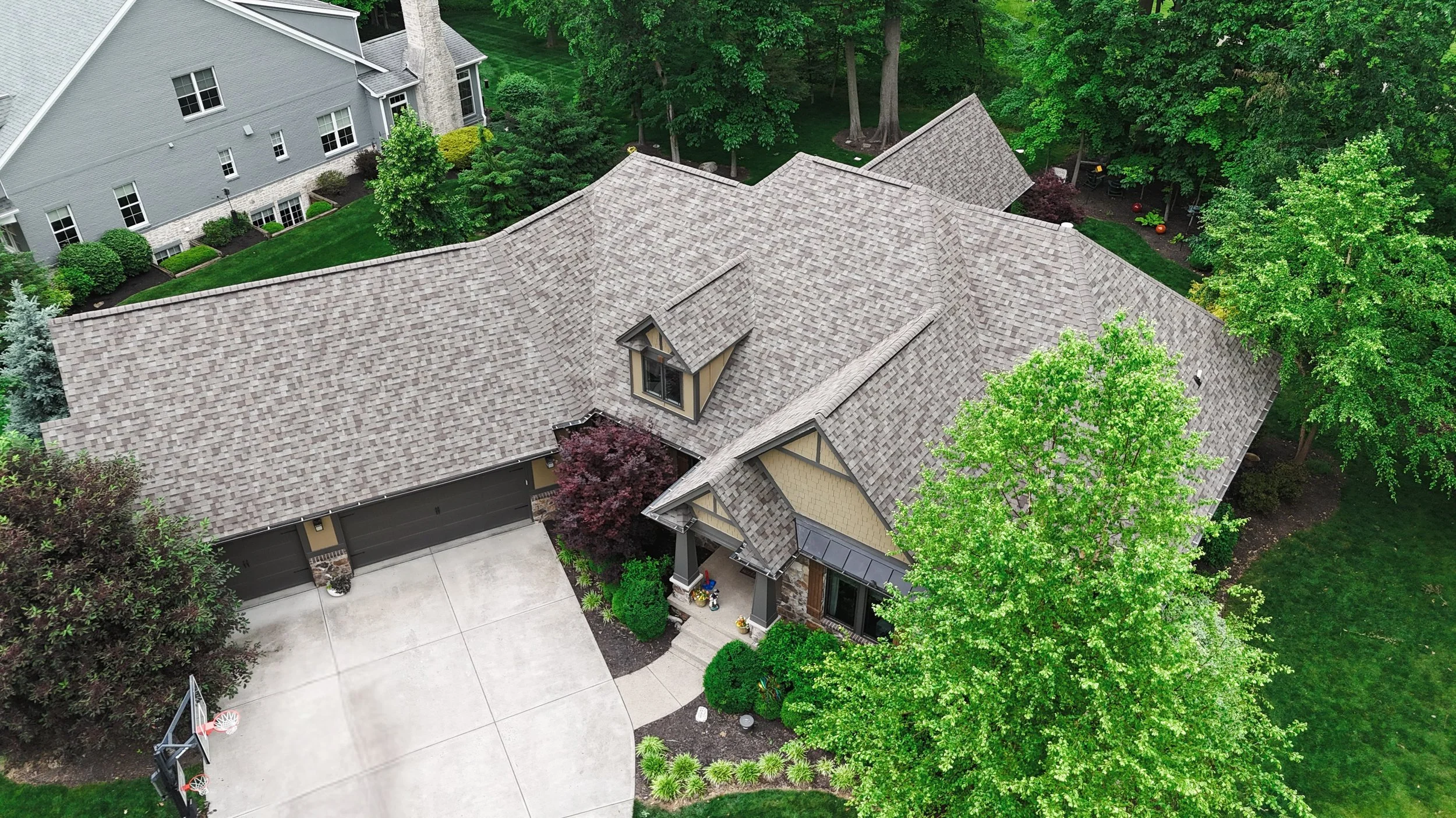 Aerial view of a suburban house with a large concrete driveway, two-car garage, grey shingle roof, and a green yard with mature trees and landscaped bushes.