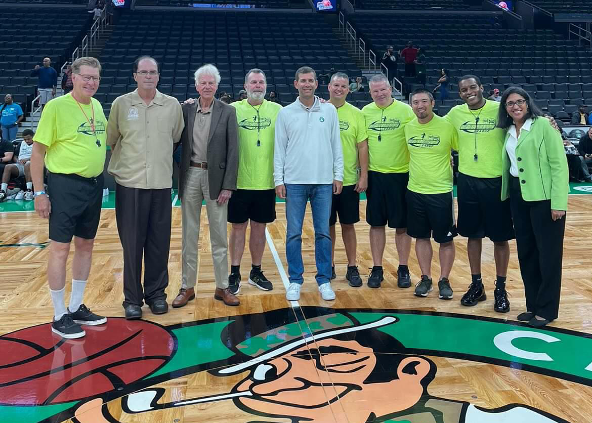 Group of ten people standing on a basketball court, smiling at the camera, with empty seats in the background. The court features a large mascot logo of a leprechaun holding a basketball.