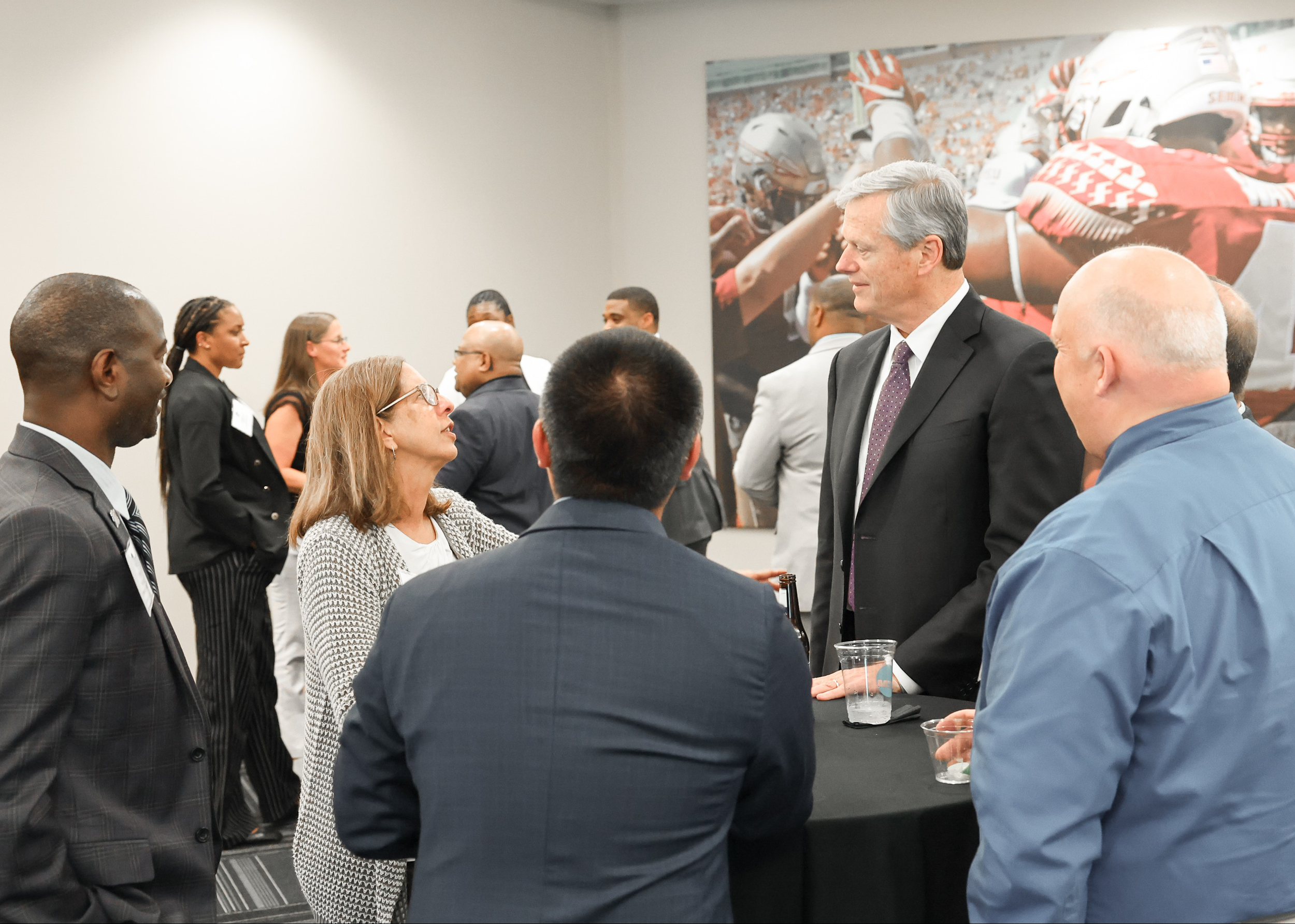 People engaged in conversation at a professional networking event, with a background poster of football players in helmets and uniforms.