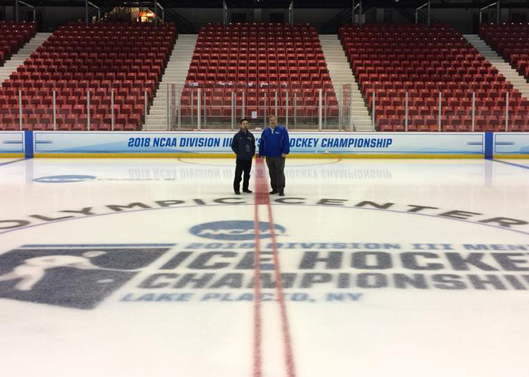 Two people standing on an ice hockey rink with empty red seats in the background and a logo on the ice for the 2018 NCAA Division III Ice Hockey Championship.