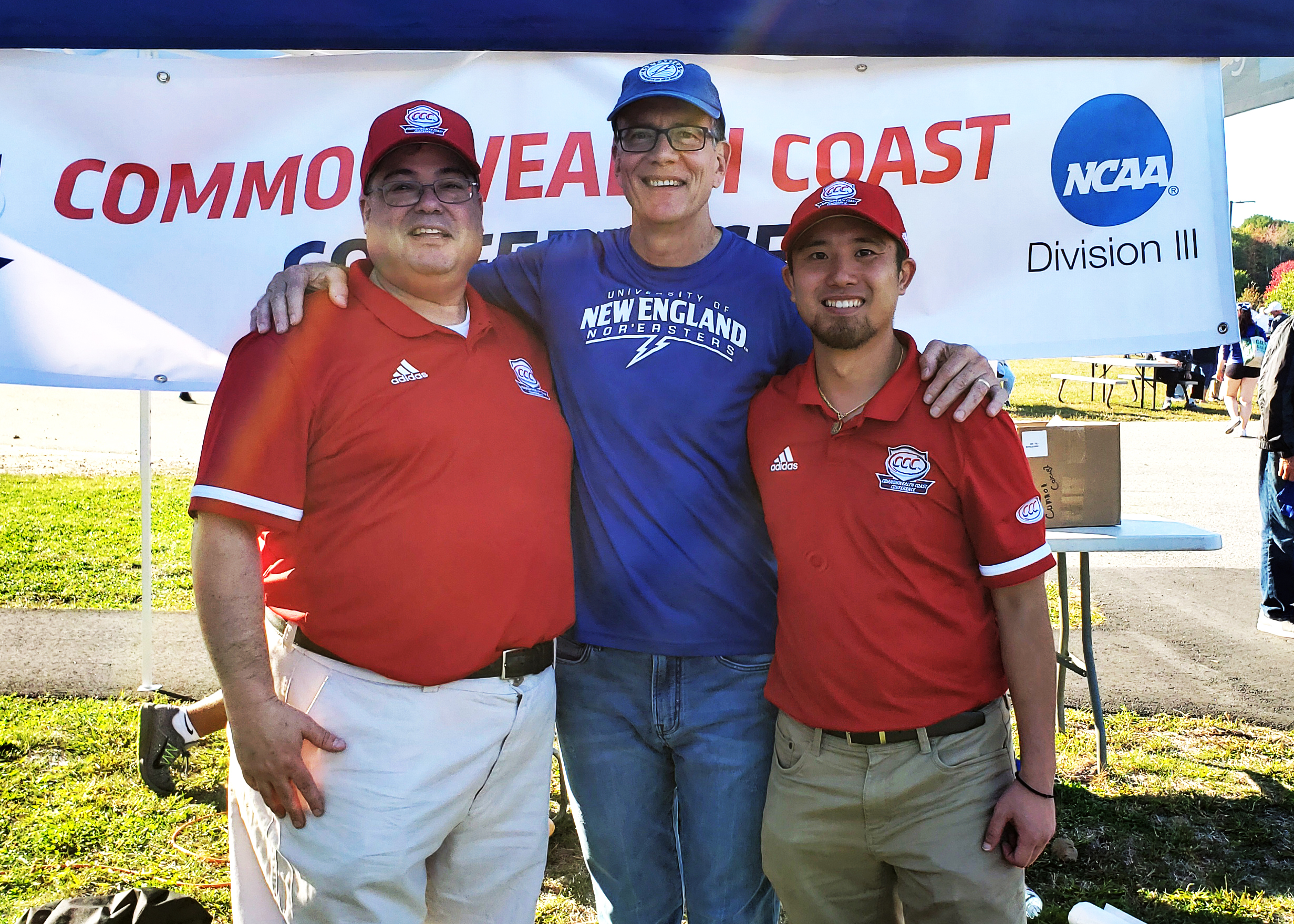 Three men standing outdoors in front of a banner that reads 'Commonwealth Coast Conference NCAA Division III.' They are wearing red and blue shirts with logos, smiling, and have their arms around each other.