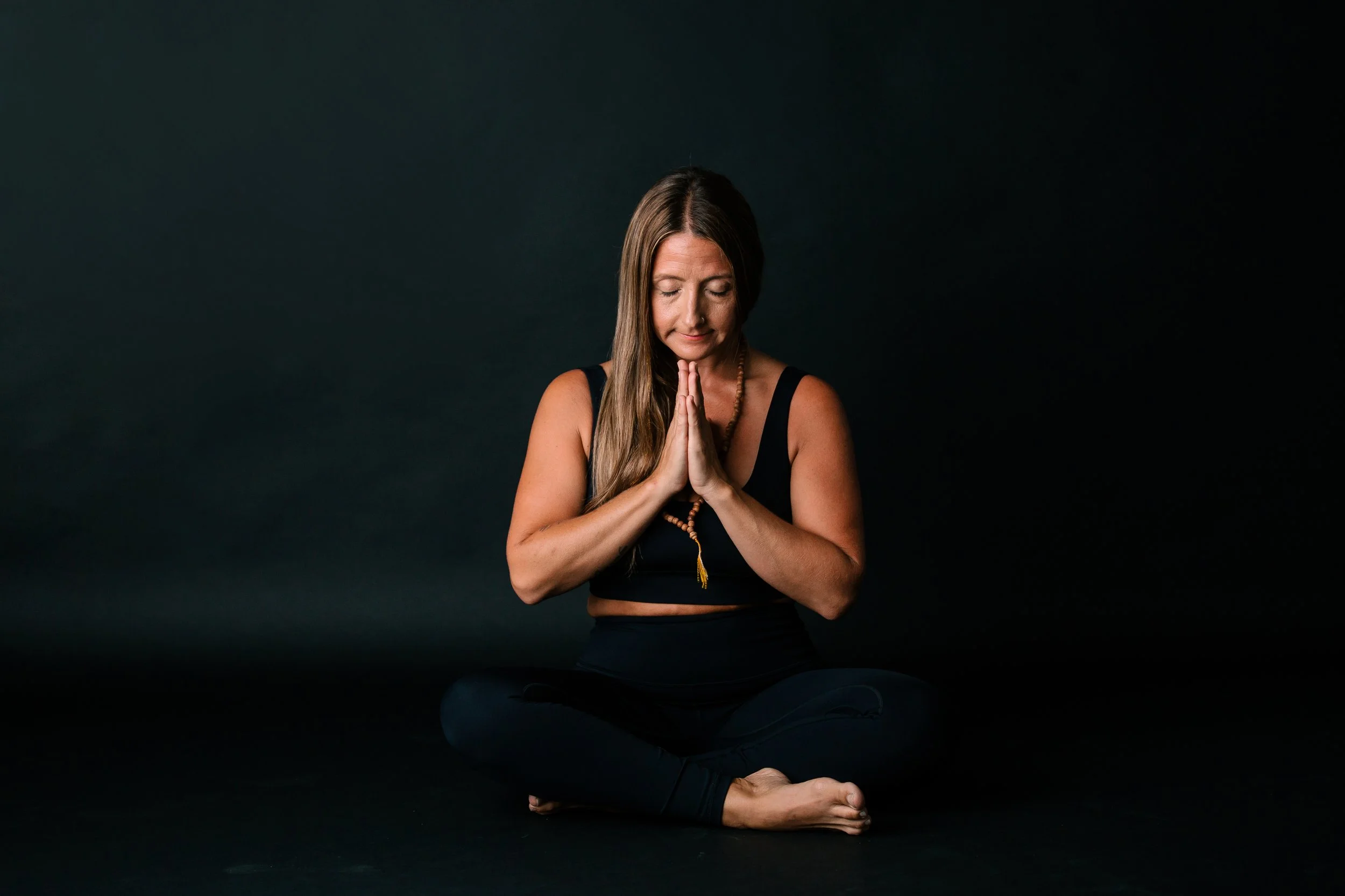 Woman in a black tank top and beaded necklace practicing yoga in a seated meditative pose on a black background, with hands pressed together in front of her chest and eyes closed.