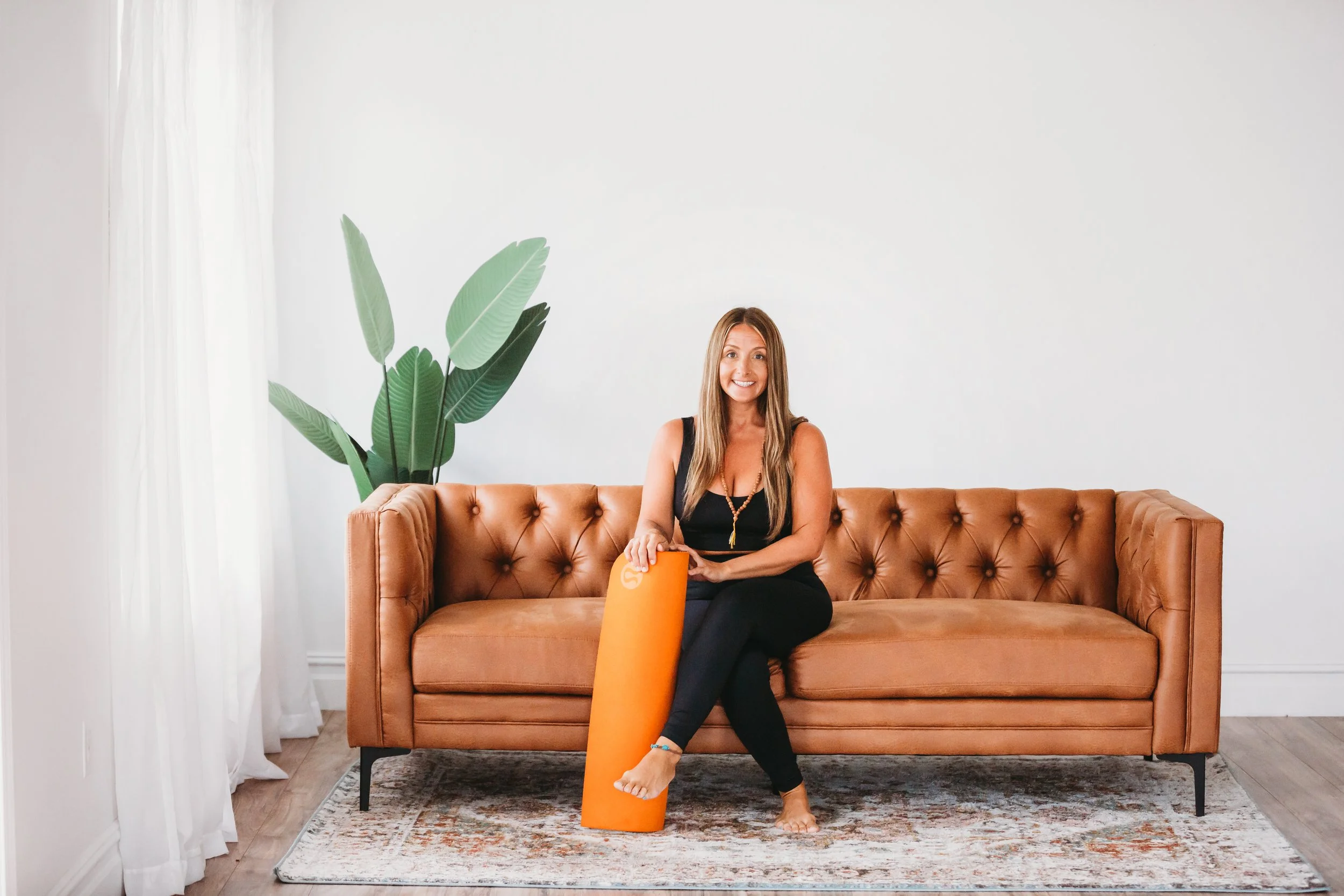 A woman sitting on a tan leather couch, smiling, with one foot resting on an orange foam roller, in a bright room with white walls, a large plant, and a patterned area rug.