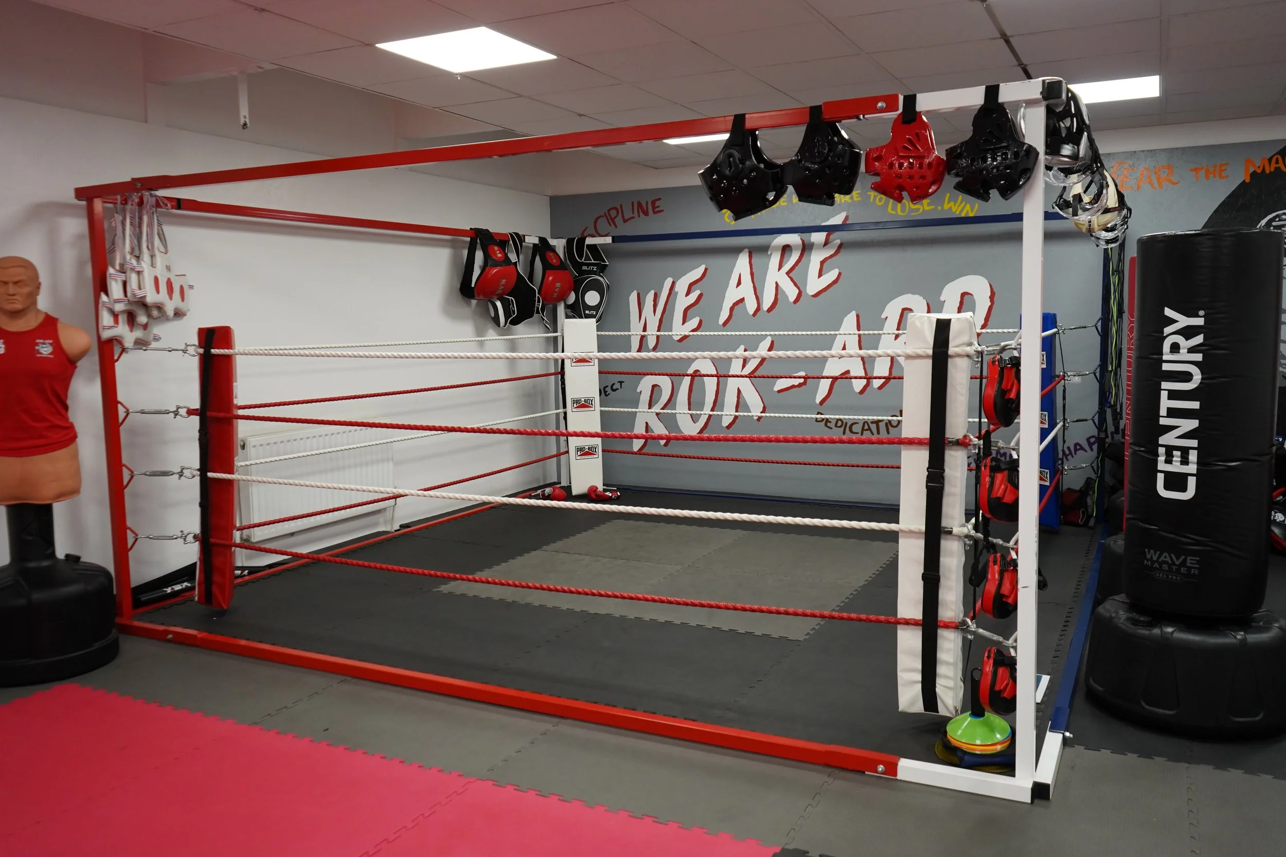 Boxing ring with red and white ropes in a gym, boxing gloves hanging on the wall, punching bags, and a mannequin torso in a red shirt on a stand.