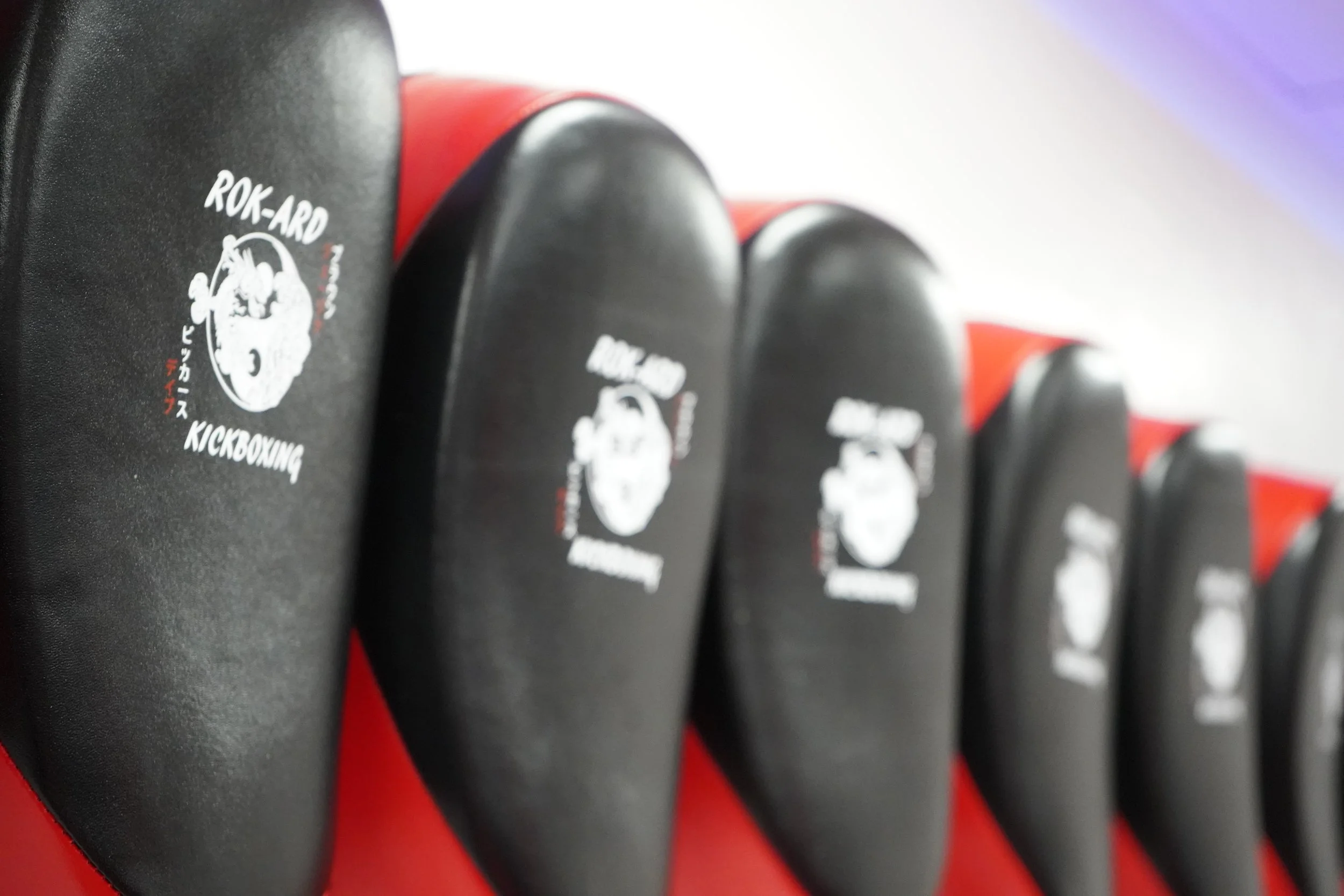 Row of black kickboxing mats with a logo that reads 'ROK-ARD Kickboxing' and Japanese characters, lined up along a red wall.