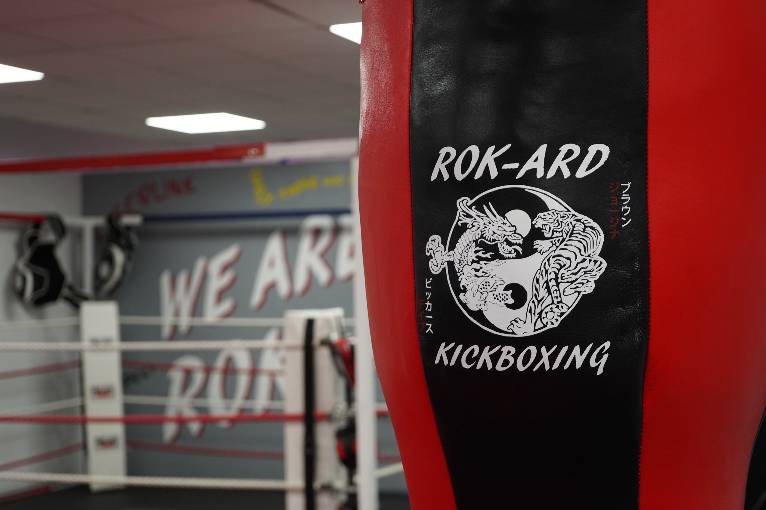 A close-up of a black and red kickboxing punching bag with a dragon illustration and the words 'ROK-ARD KICKBOXING' on it, inside a boxing gym with a boxing ring and a wall with the phrase 'WE ARE' visible in the background.