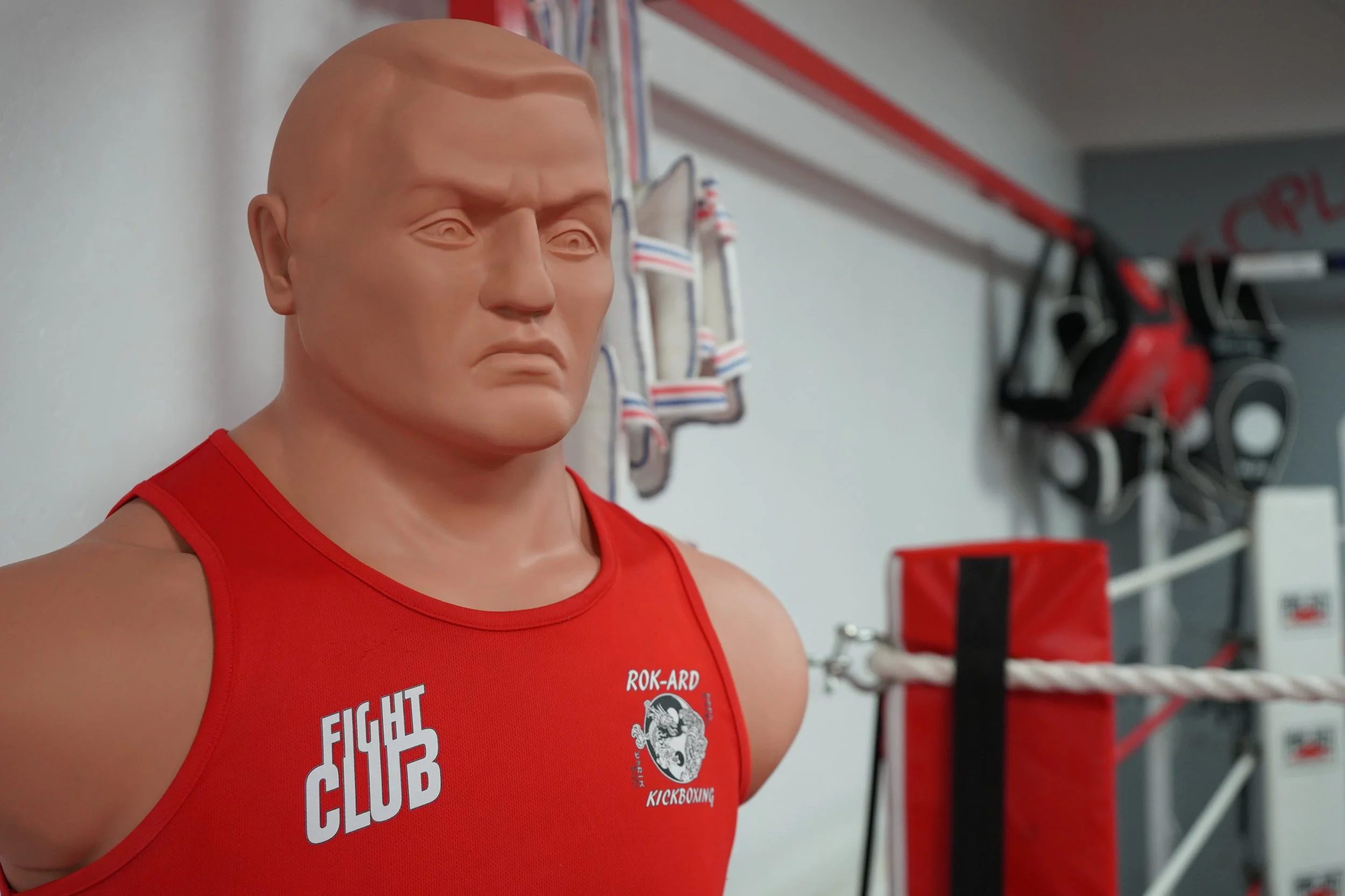 A human-like training dummy with a serious expression, wearing a red tank top with the words 'FIGHT CLUB' and 'ROK-ARD KICKBOXING' on it, inside a gym with boxing equipment and punching bags in the background.