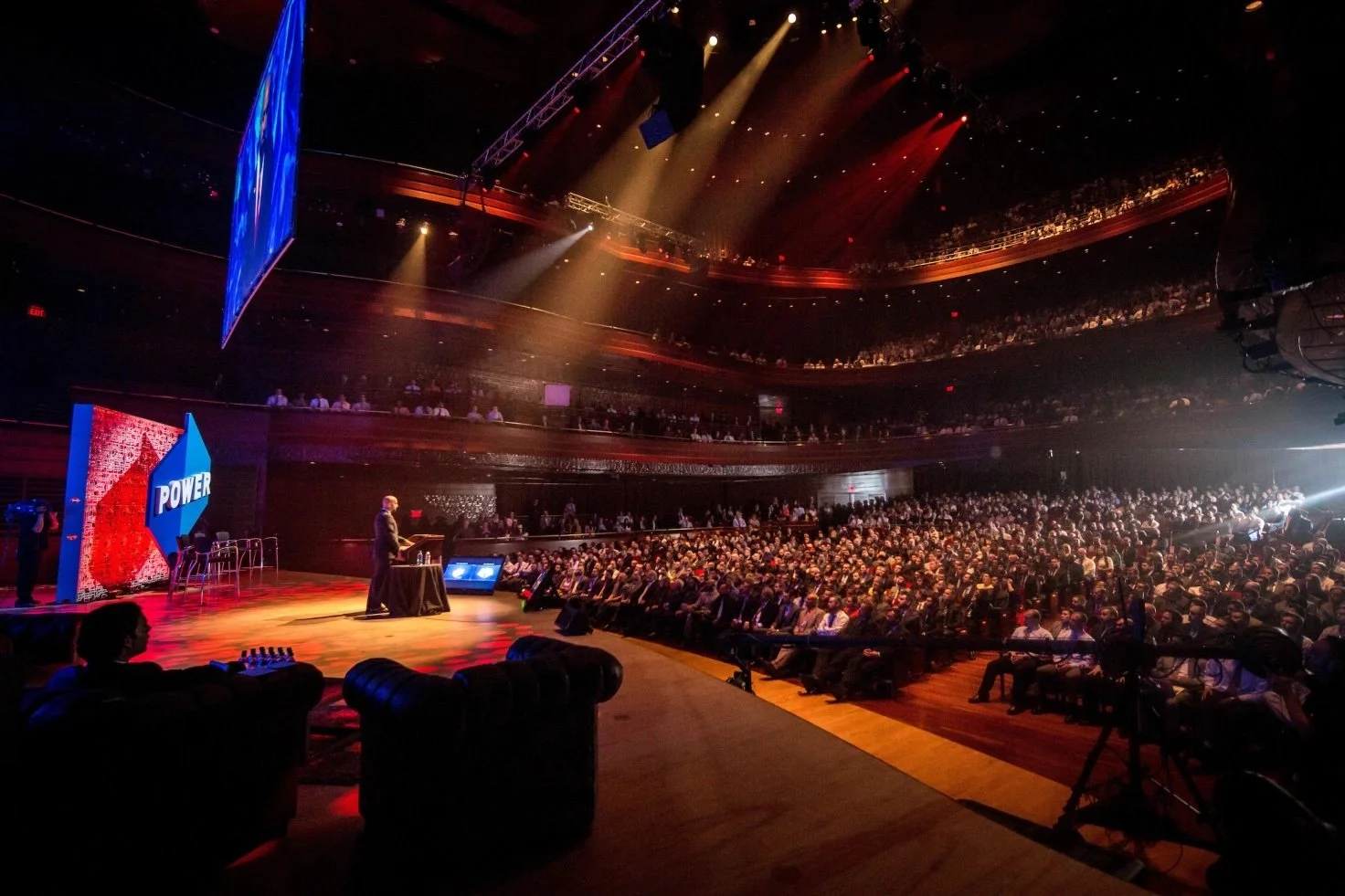 Large auditorium filled with people attending a conference or presentation. A stage with a speaker and large screens displaying the word 'POWER' is visible. The audience is seated in multiple tiers, and stage lighting illuminates the scene.