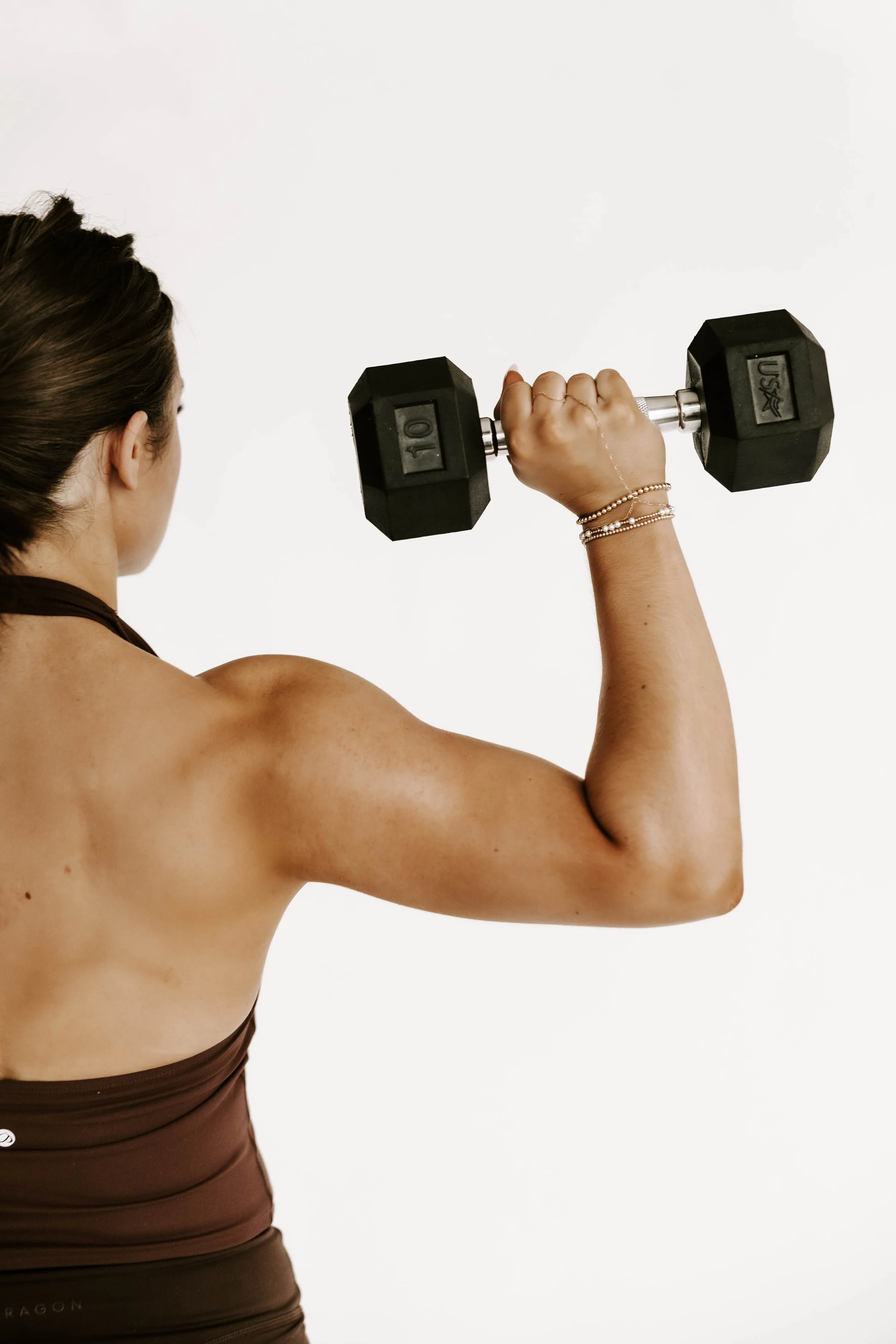 A woman lifting a black dumbbell with her right arm, wearing a brown sports top and jewelry, against a plain white background.