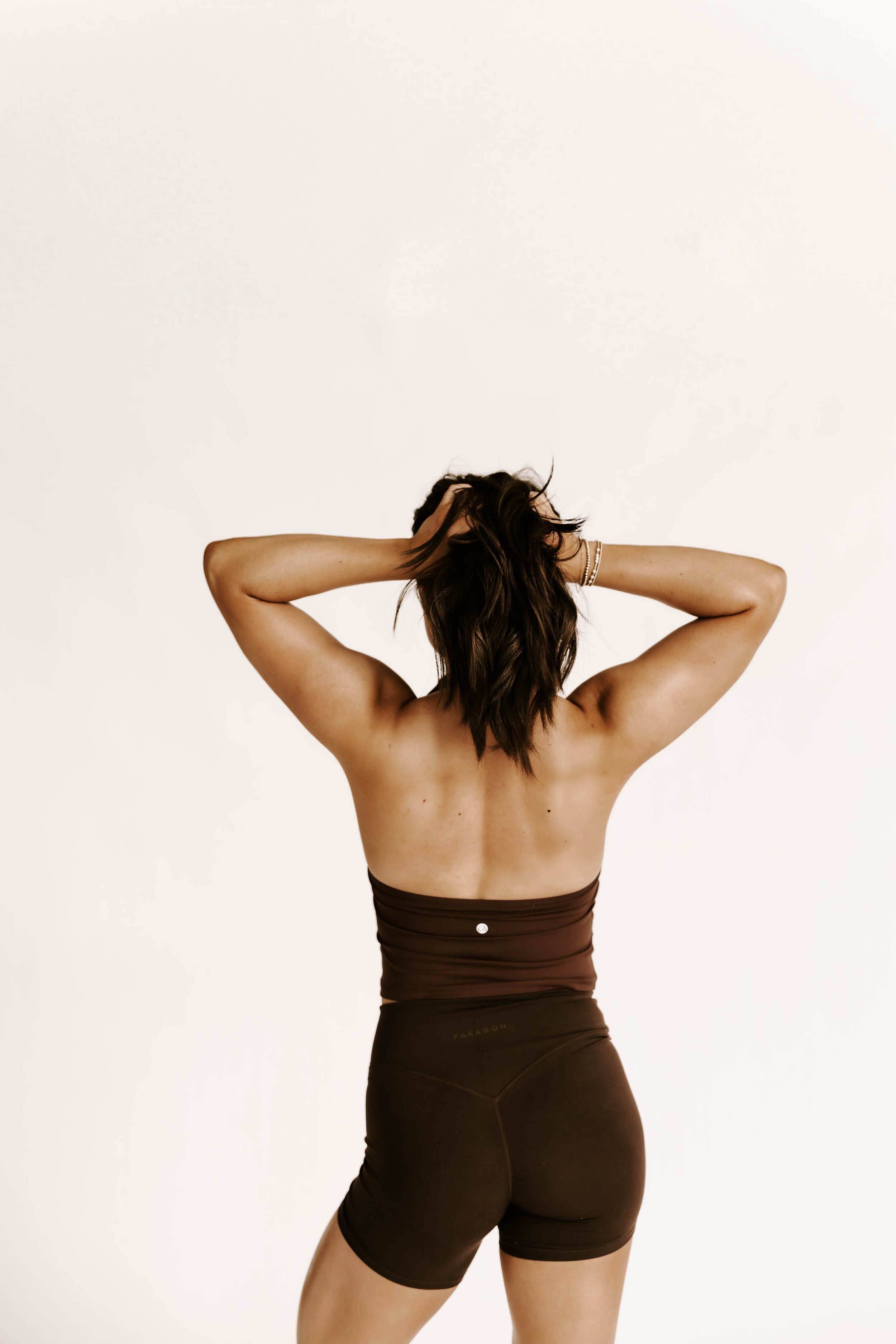Woman in athletic wear with her back to the camera, hands in her hair, against a plain white background.