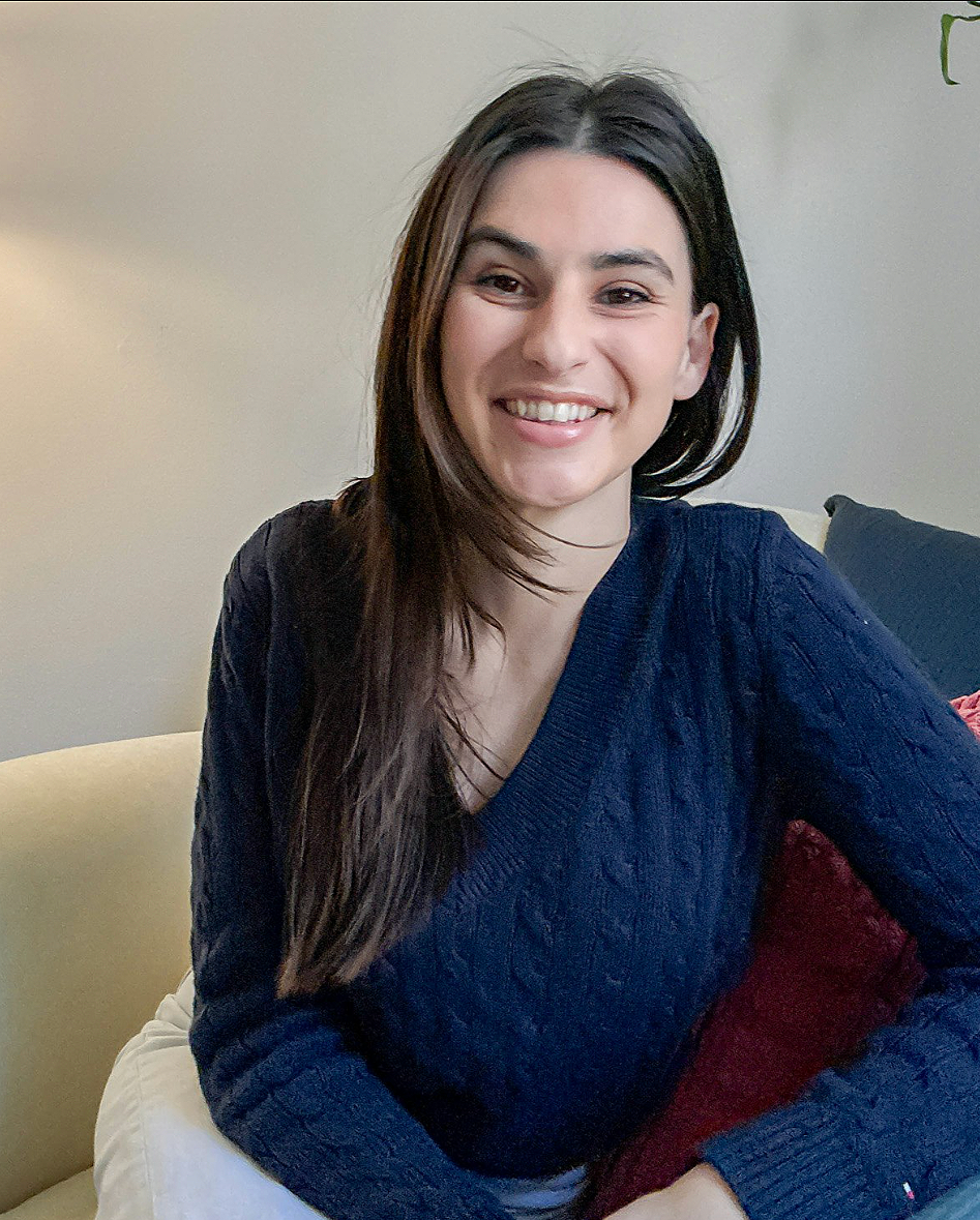 A young woman with long dark hair smiling, sitting on a beige and red couch, wearing a navy blue sweater.