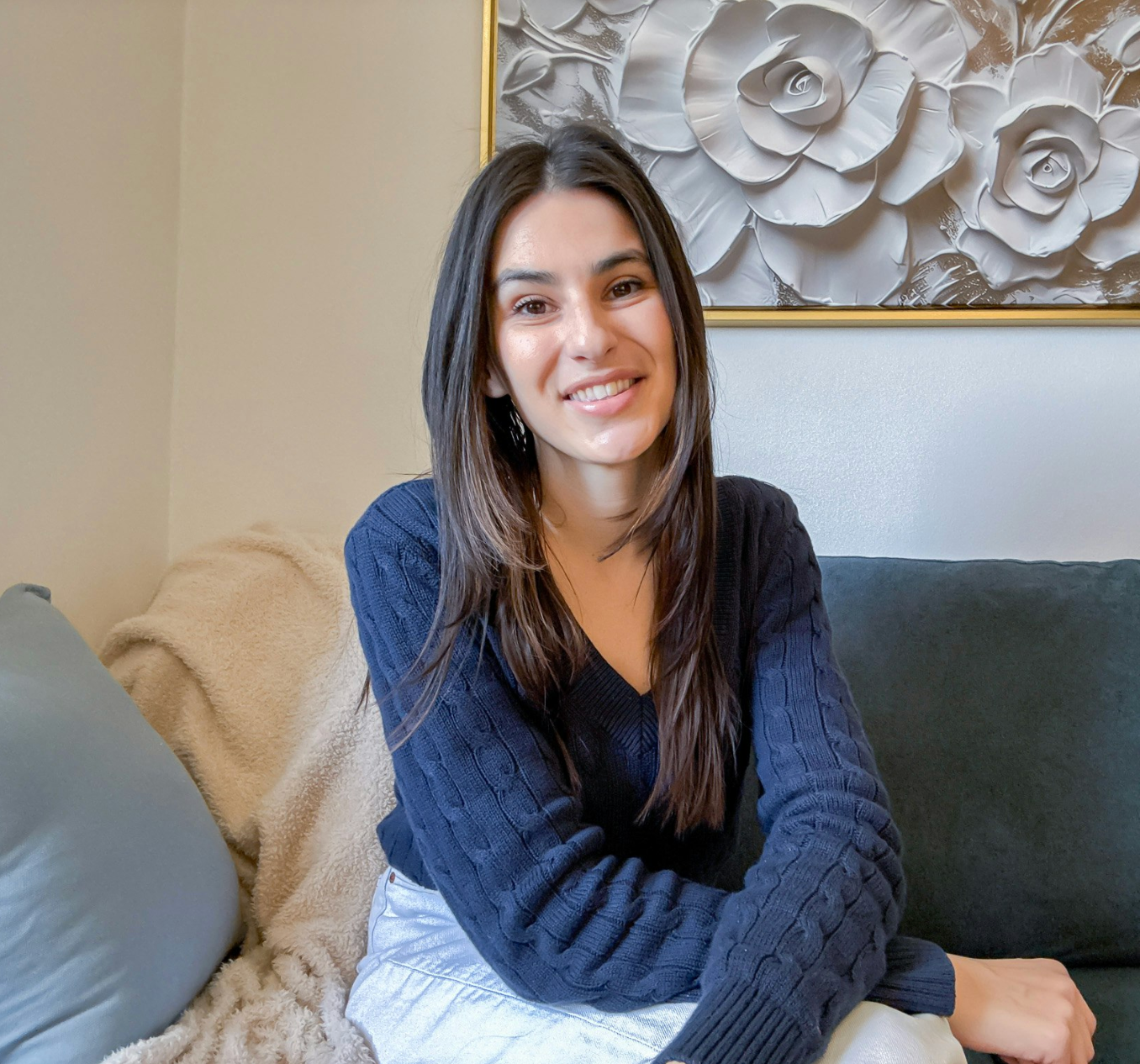 A young woman with long brown hair, smiling and wearing a navy blue sweater, sitting on a couch with a beige blanket and gray pillows, in a room with a floral painting on the wall.