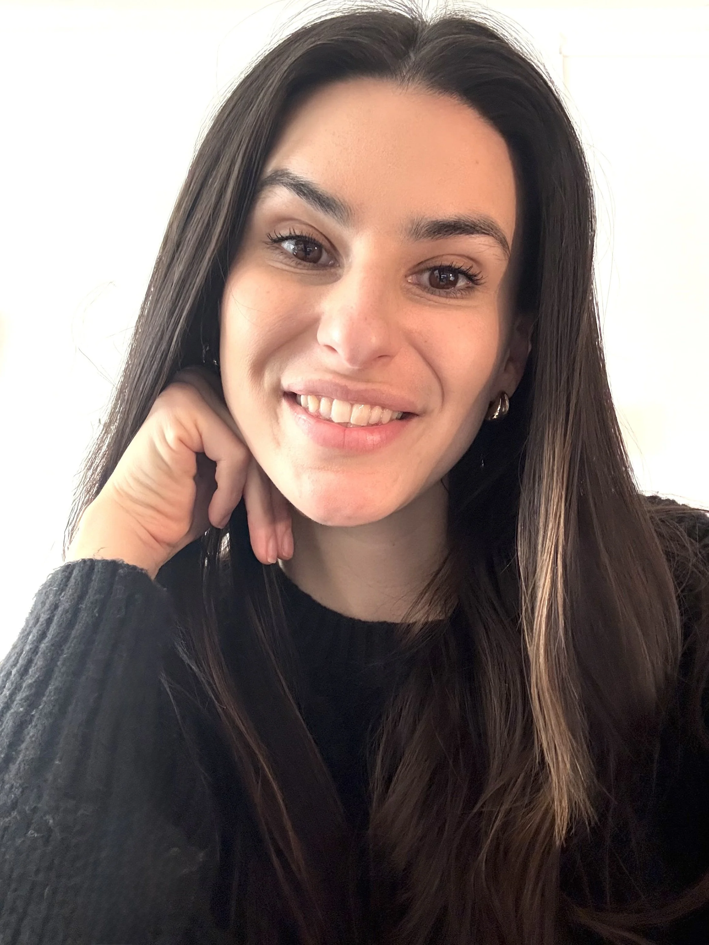 Close-up of a young woman with long dark hair smiling and resting her chin on her hand, wearing a black sweater and small hoop earrings, against a light background.