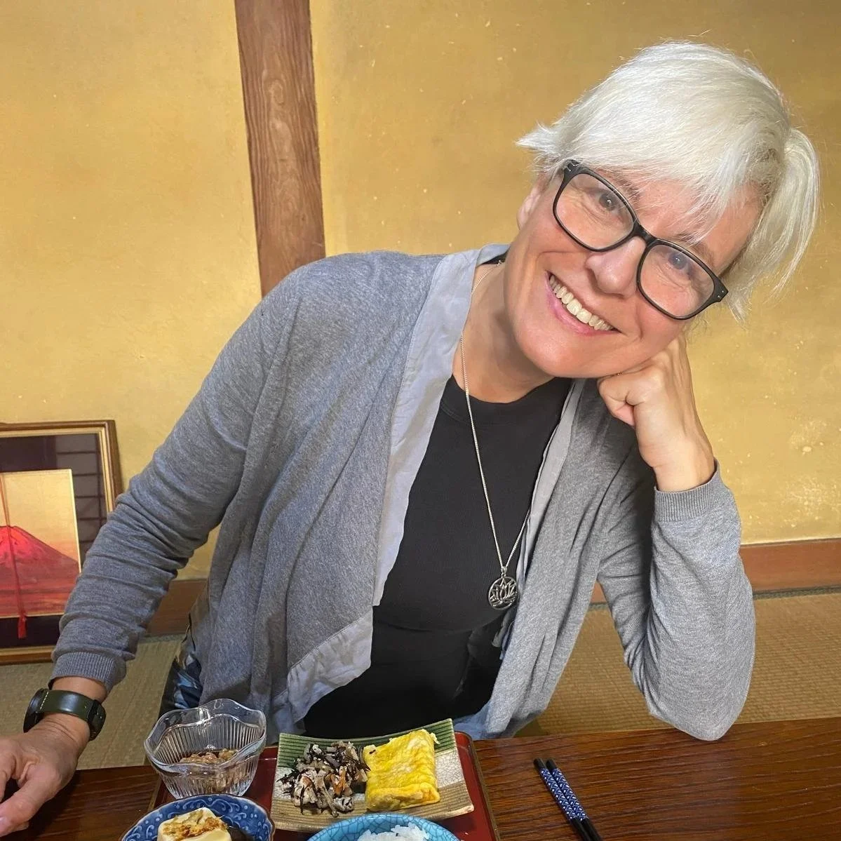 A smiling woman with short gray hair and glasses sitting at a table with Japanese food, resting her head on her hand.