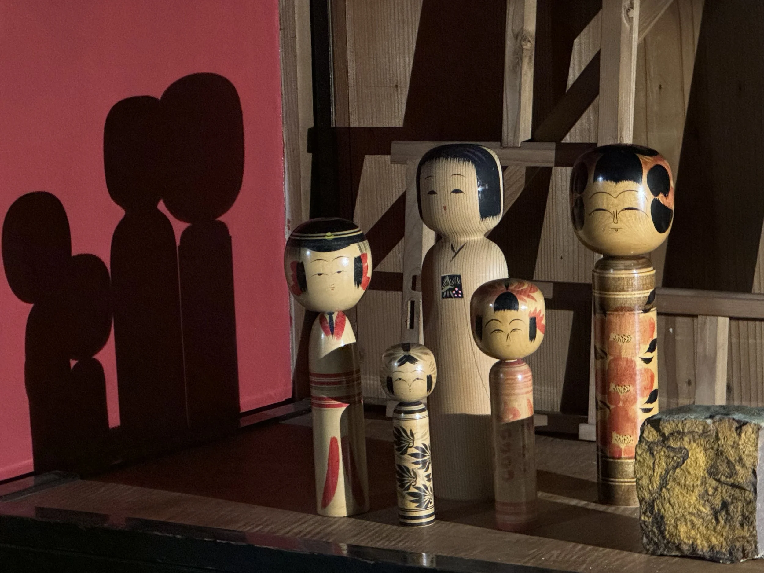 Set of six traditional Japanese nesting dolls with smiling faces, decorated with floral and geometric patterns, placed on a wooden surface with a stone nearby. Shadows cast on a red wall behind them.