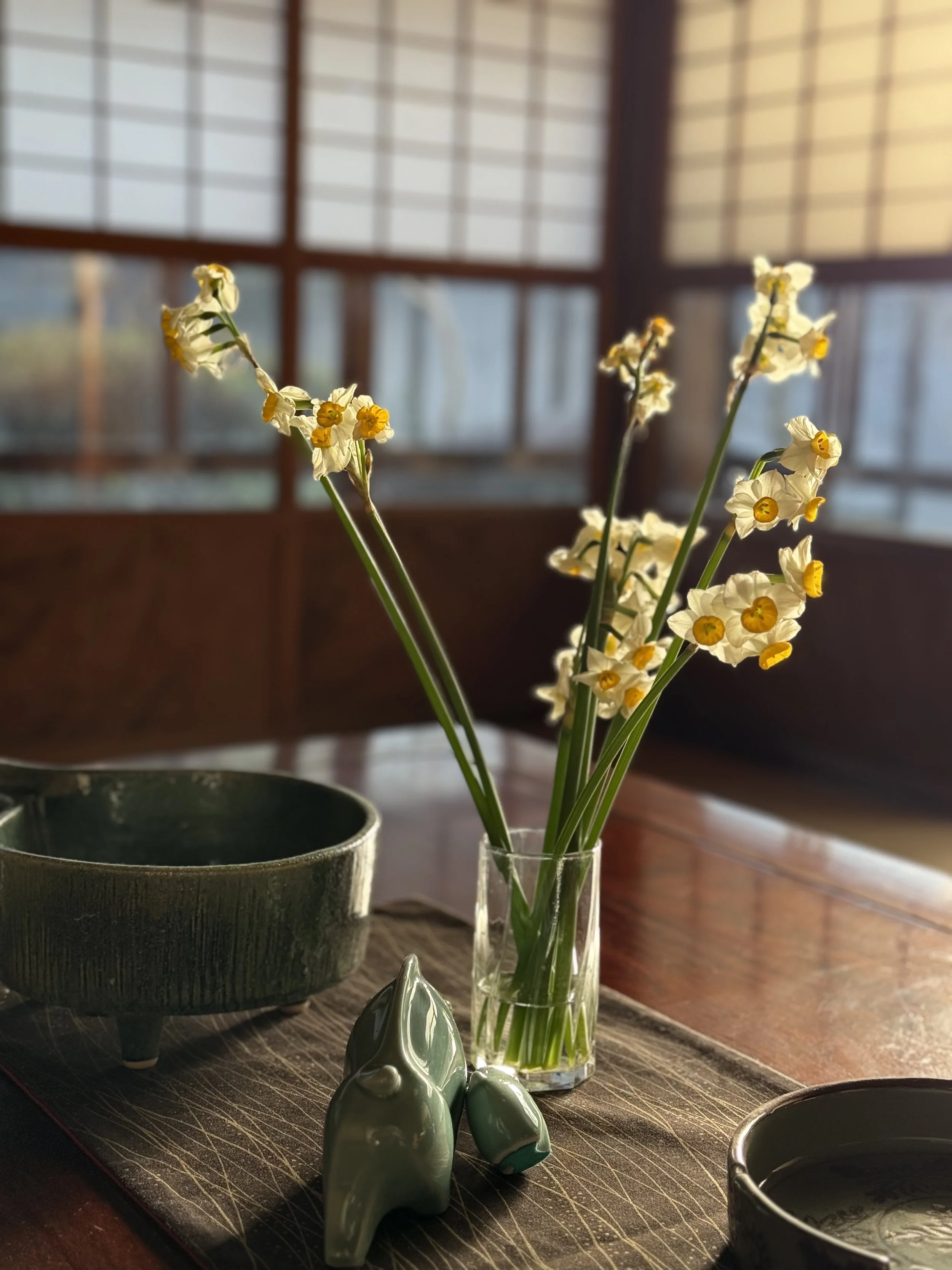 A small vase with white and yellow flowers on a wooden table, with a ceramic bowl and a ceramic frog figurine nearby, and shoji-style window in the background closing the room with soft natural light.
