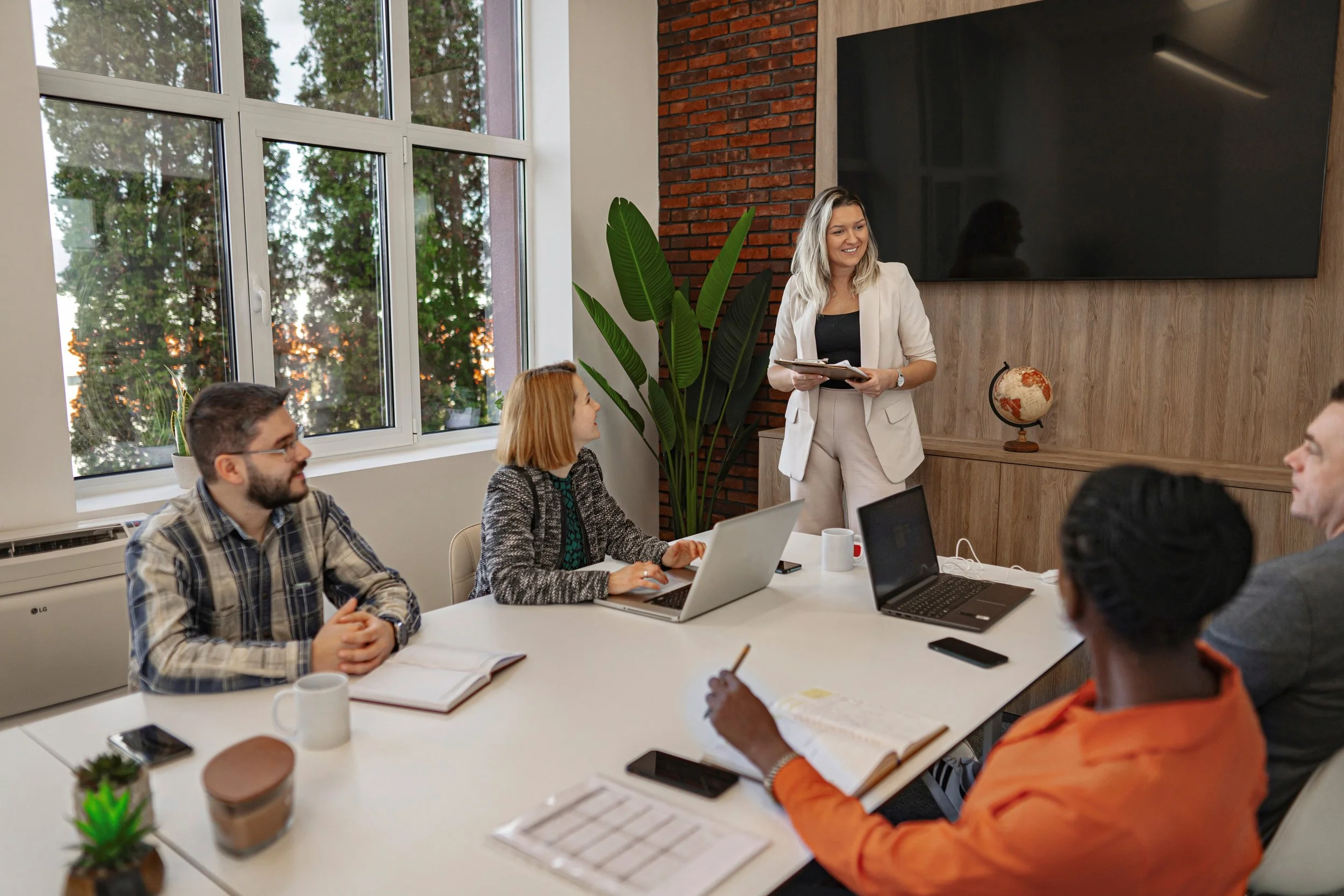 A diverse group of five colleagues attending a meeting in a modern office. A woman standing and smiling holds a clipboard, while four others sitting at a white table with laptops, notebooks, and cups listen attentively. Large windows reveal trees outside, and a flat screen and a globe are on the wall behind.
