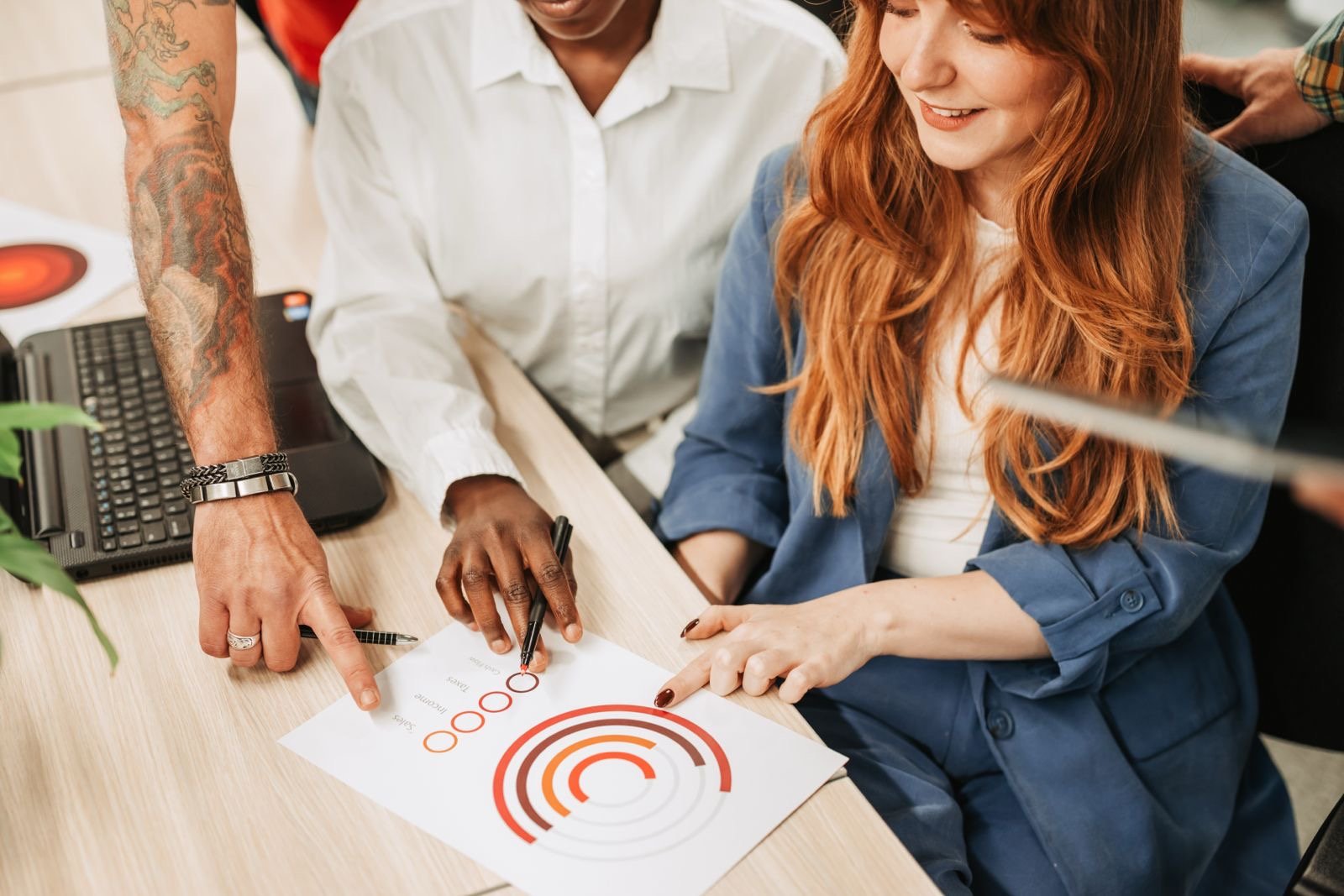 Two people at a desk, one with tattooed arm, pointing at a chart with circles and concentric arcs, while the other woman watches, smiling.