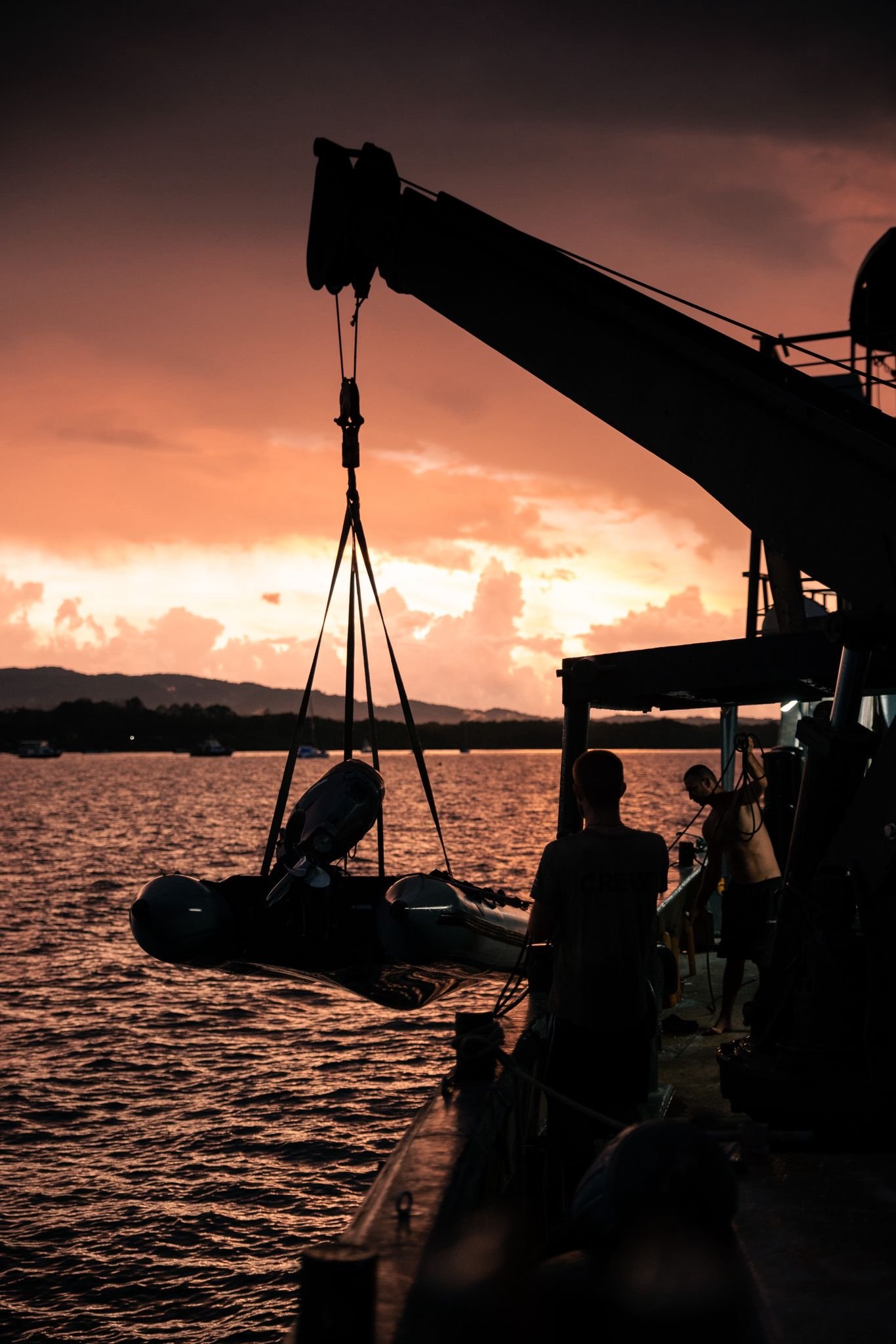Silhouette of a boat crane on conservation vessel Modoc lifting a small boat/Zodiach at sunset, with two crew/volunteers on the boat deck, over a body of water with hills in the distance.