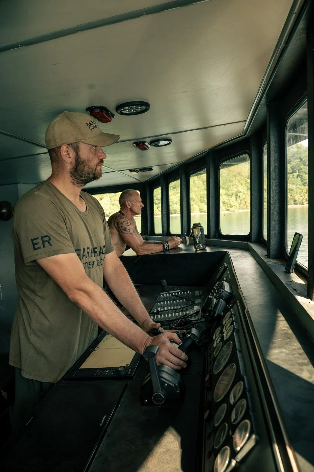 Two men (crew from NGO Earthrace Conservation in Costa Rica) in a boat's control room called bridge, one operating the controls and the other sitting by the windows overlooking water and trees. The conservation vessel is called Modoc, ex-navy vessel