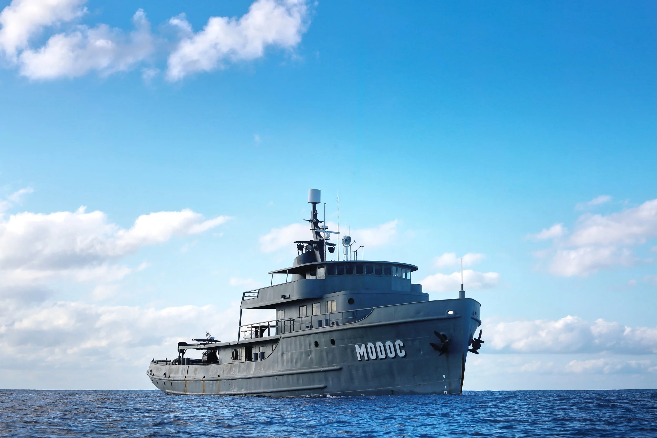 A gray naval ship named MODOC floating on the ocean under a blue sky with scattered clouds.