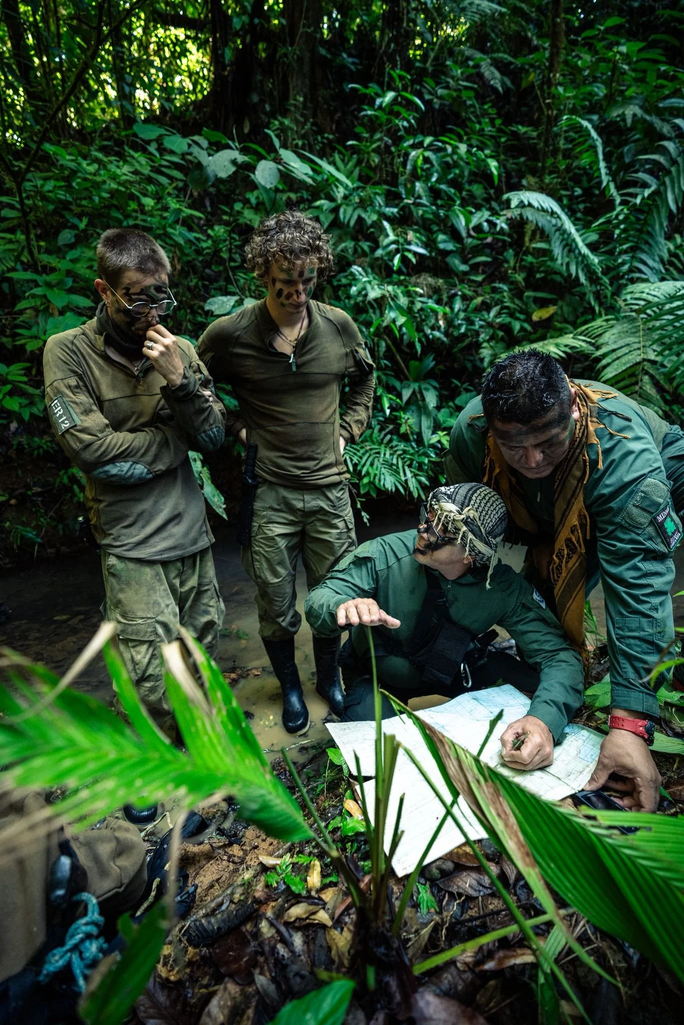 Group of four people in military or expedition gear, some with face paint, gathered in dense jungle. One person is sitting on the ground pointing to a map, while others stand nearby observing the discussion, surrounded by thick green foliage.