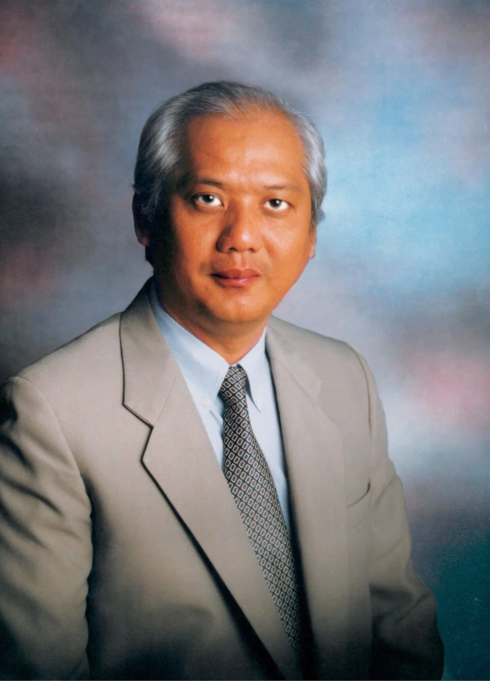 Formal portrait of a middle-aged Asian man with gray hair wearing a beige suit, light blue shirt, and patterned tie