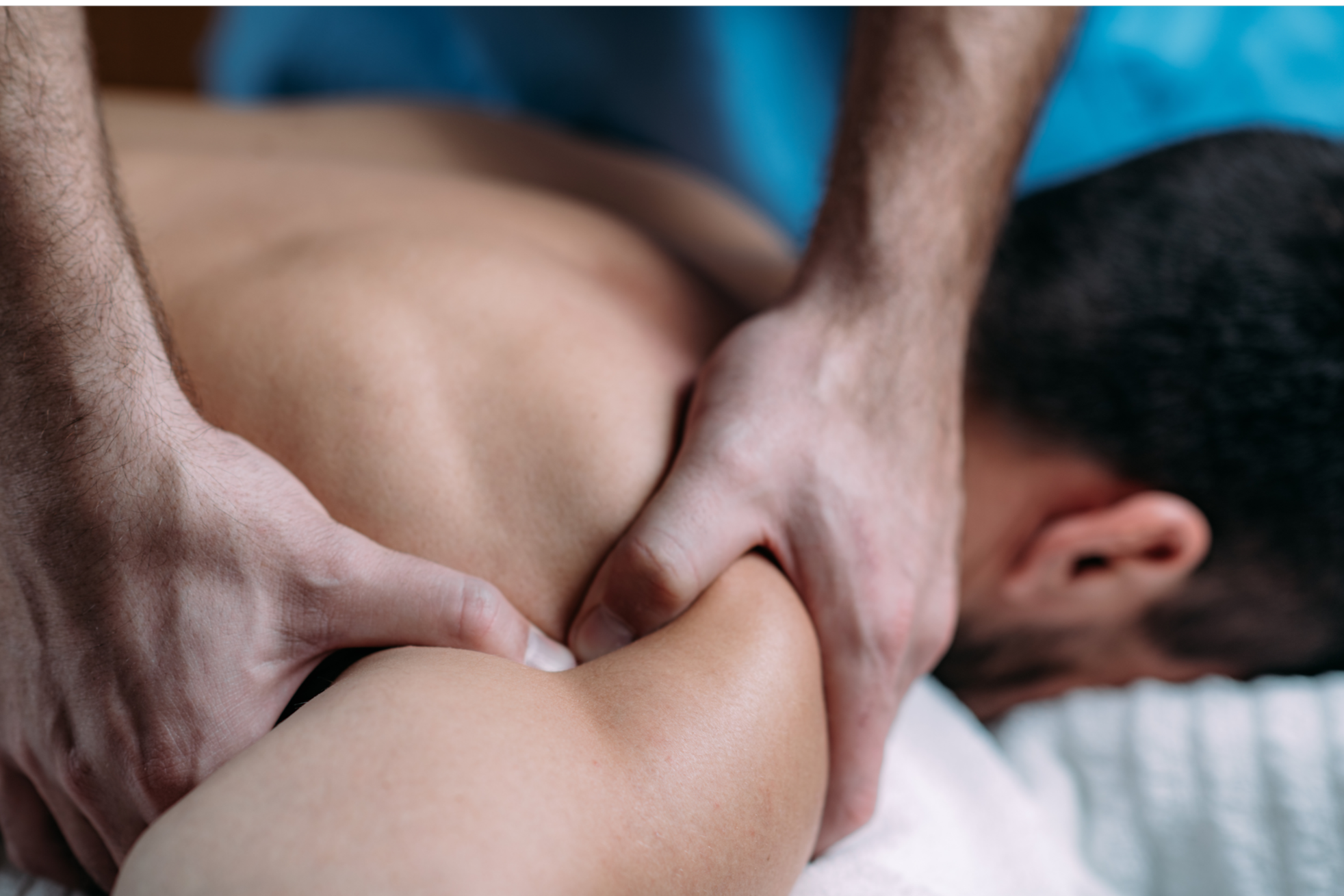 An image of a male patient face down on a massage table.  A body work provider is using their hands to apply pressure to his right shoulder to release tension