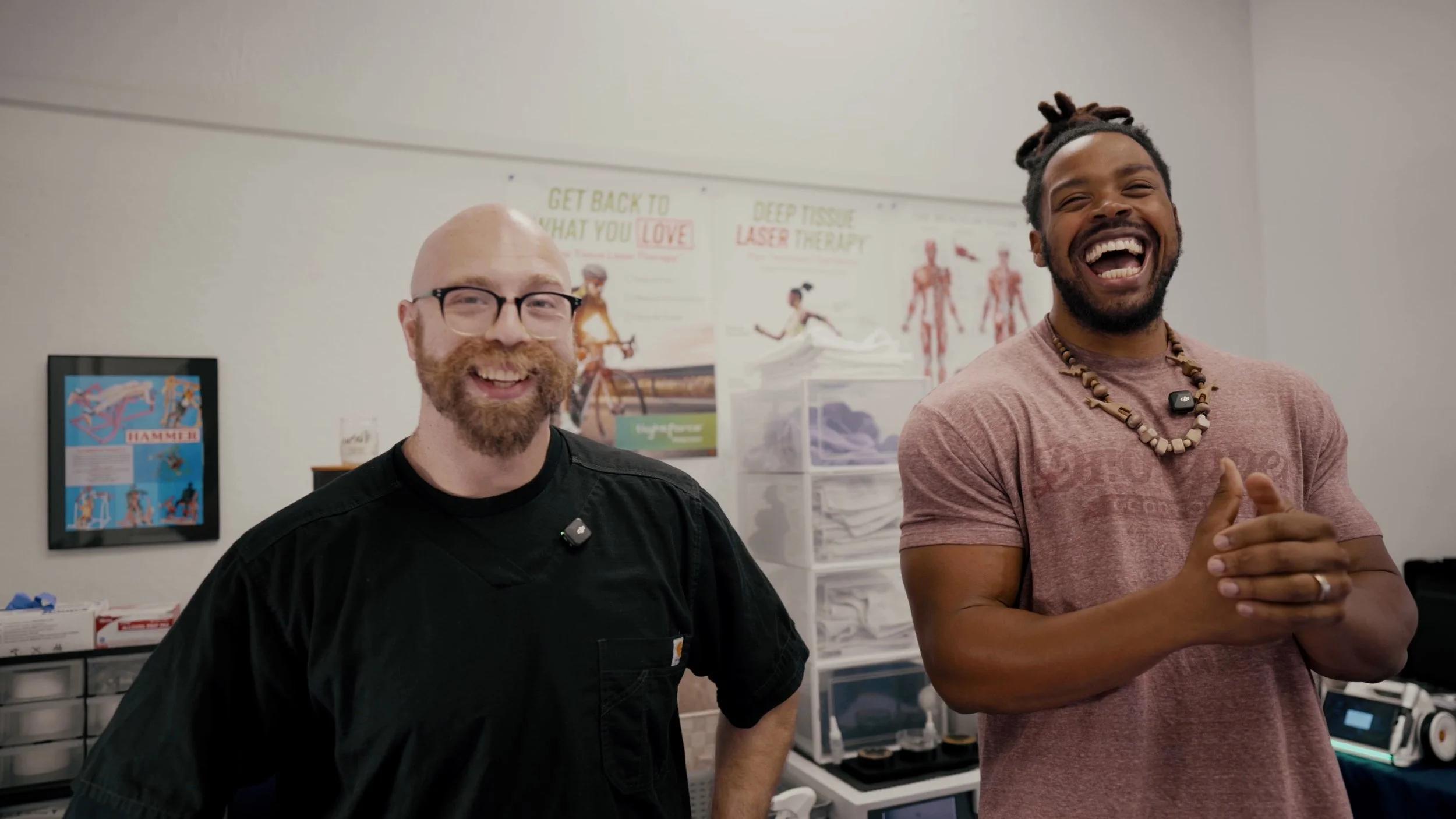 Two men smiling and laughing indoors, with medical posters and supplies in the background.
