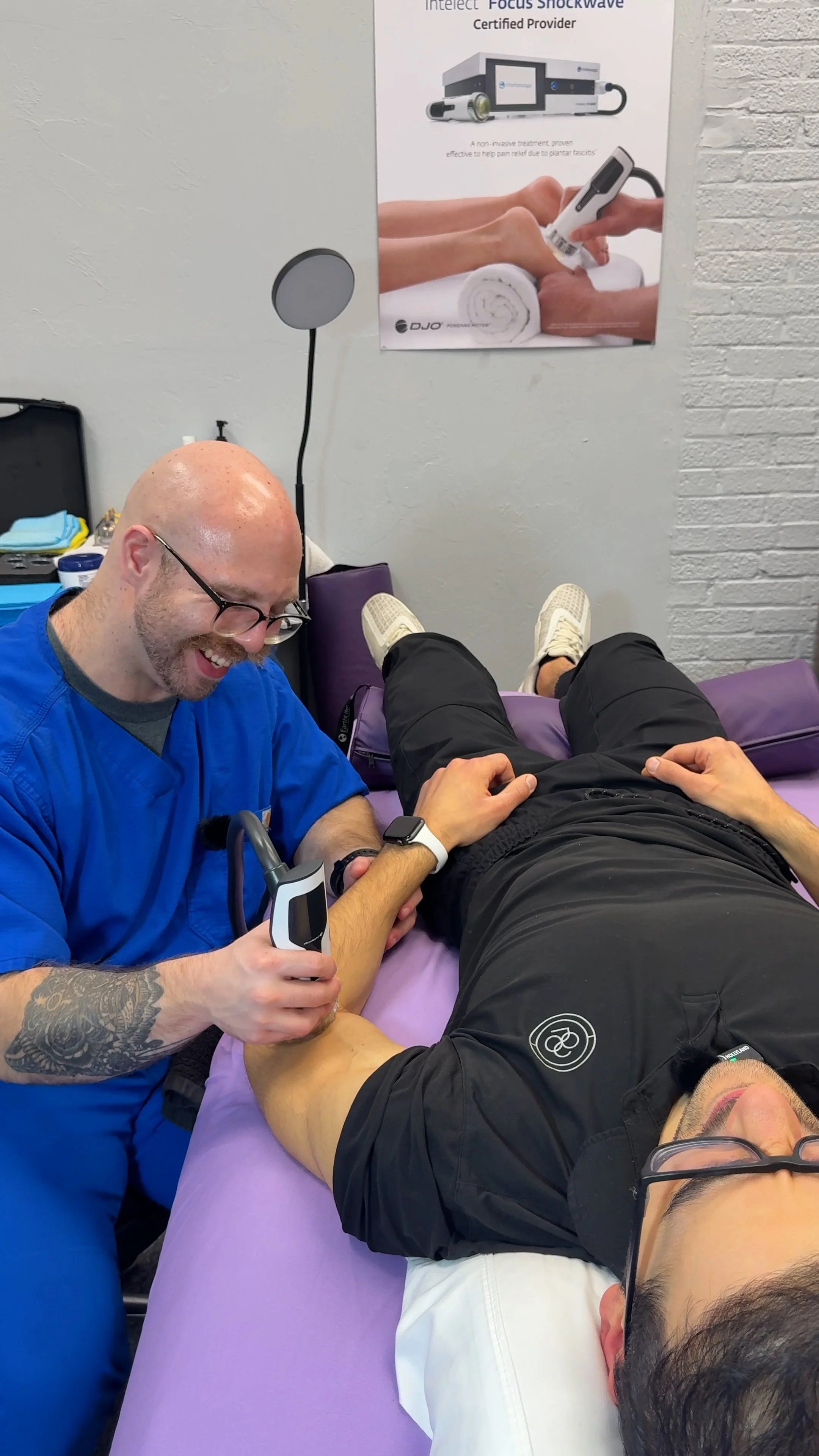 A healthcare professional using a handheld medical device on a patient's knee during a therapy session in a clinic, with a poster on the wall about shockwave therapy.