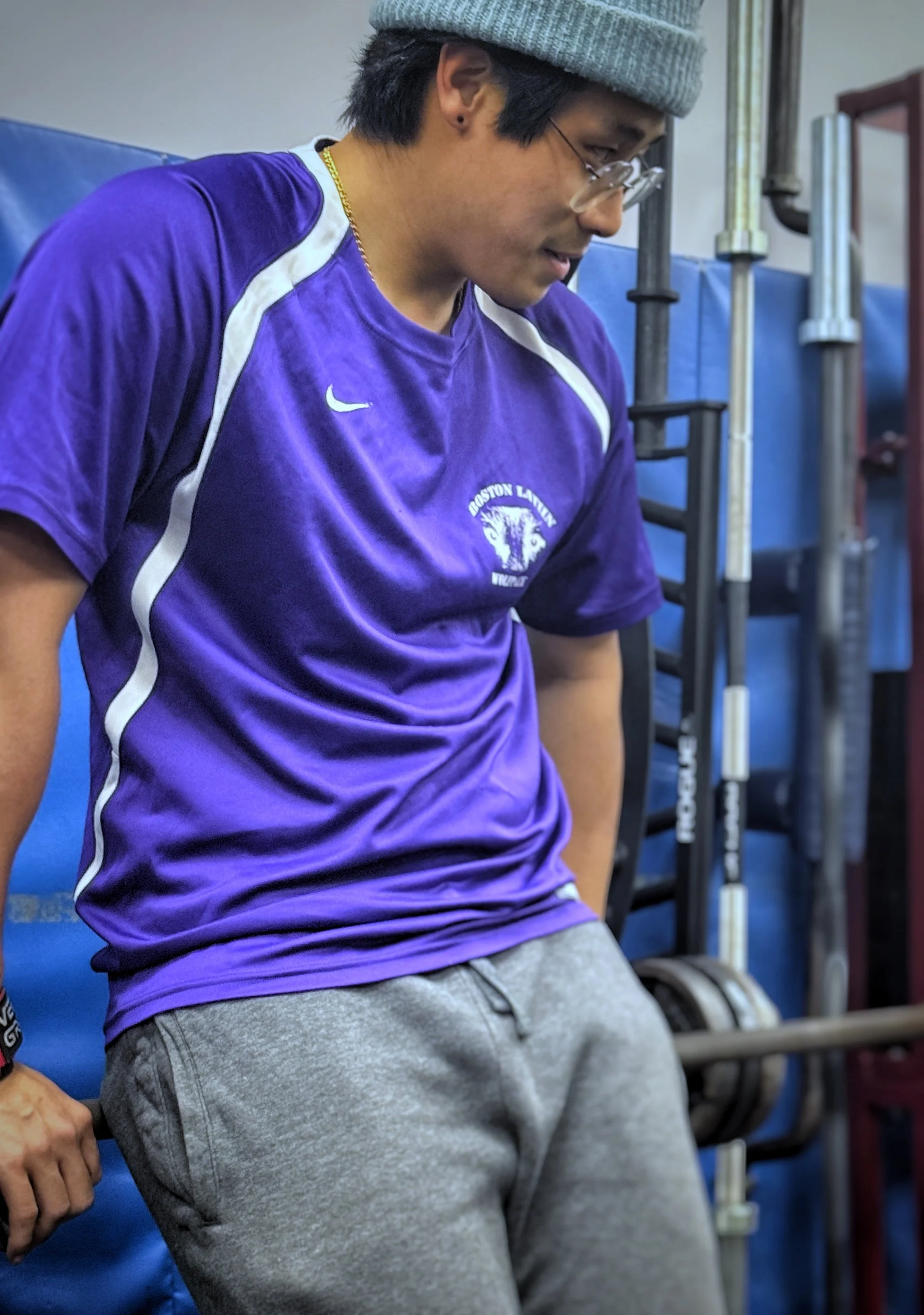 A man wearing glasses, a gray beanie, a purple sports shirt with white accents and the logo of Boston Latin Academy, gray sweatpants, standing in a gym or exercise area, leaning against a barbell.
