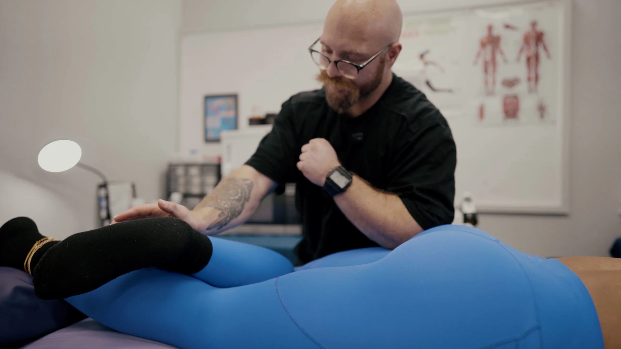 A male physical therapist with glasses and a beard working with a patient on a physical therapy bed. The patient is lying face down, wearing blue leggings and black socks, with their legs and lower back exposed. The therapist is examining or treating the patient's lower back, in a clinical setting with anatomical charts on the wall.