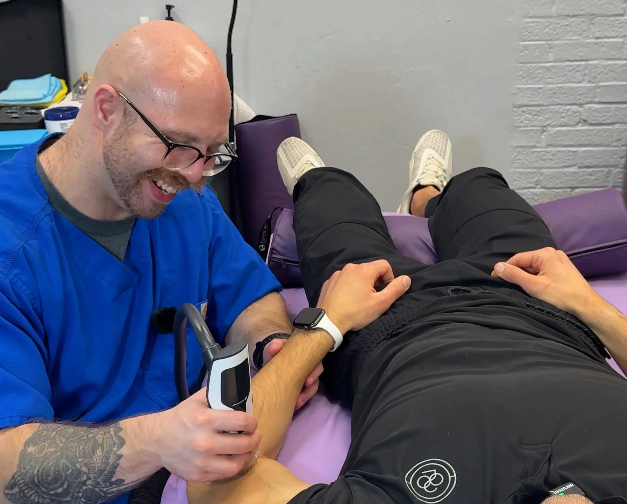 Nate Tellen smiling, wearing blue scrubs while he uses the shockwave on his client's left elbow. Client is wearing a white watch on his left wrist and a black shirt, black pants, and white shoes