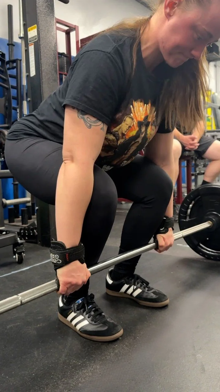 A woman with long hair, tattoos, and wearing black workout clothes and sneakers, is lifting a barbell in a gym.