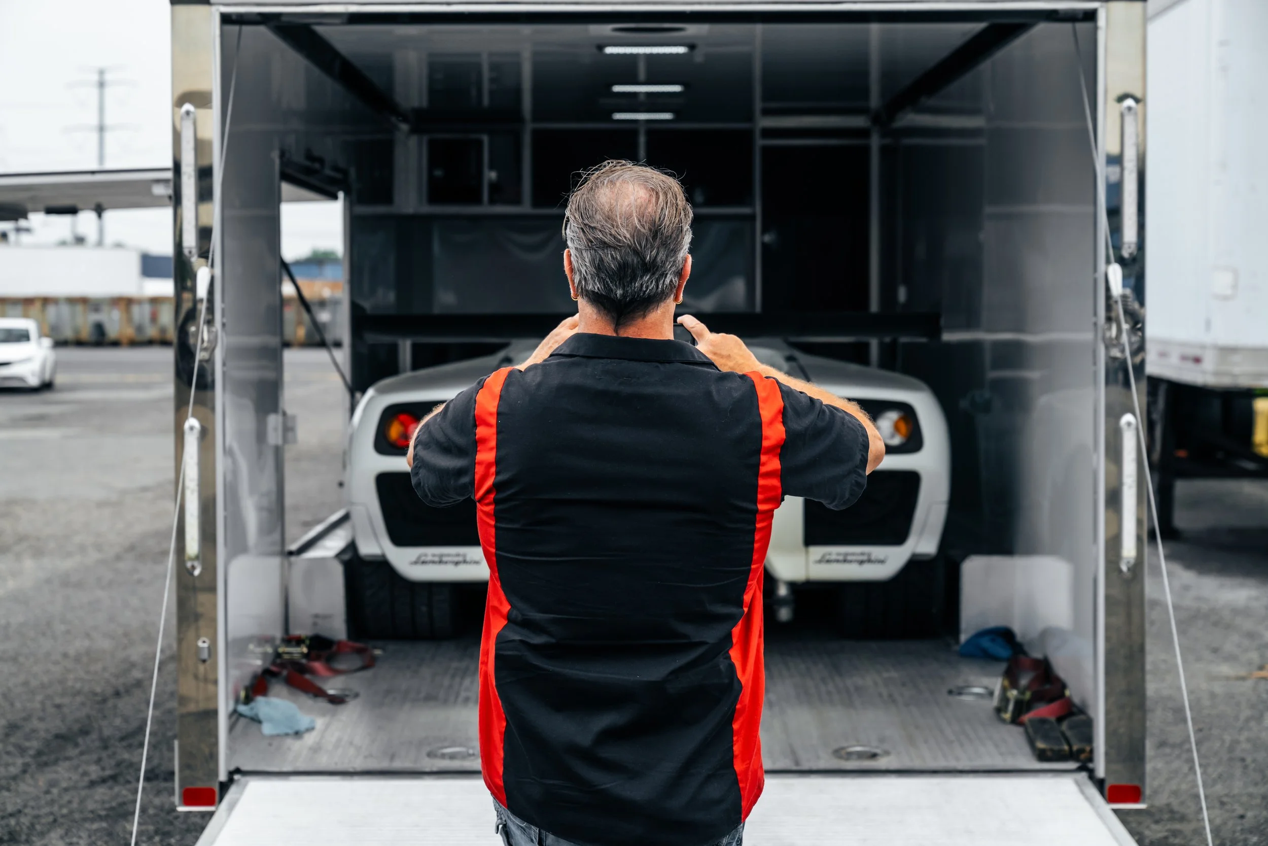 A man with gray hair wearing a black and red racing team shirt preparing a white sports car inside a trailer.