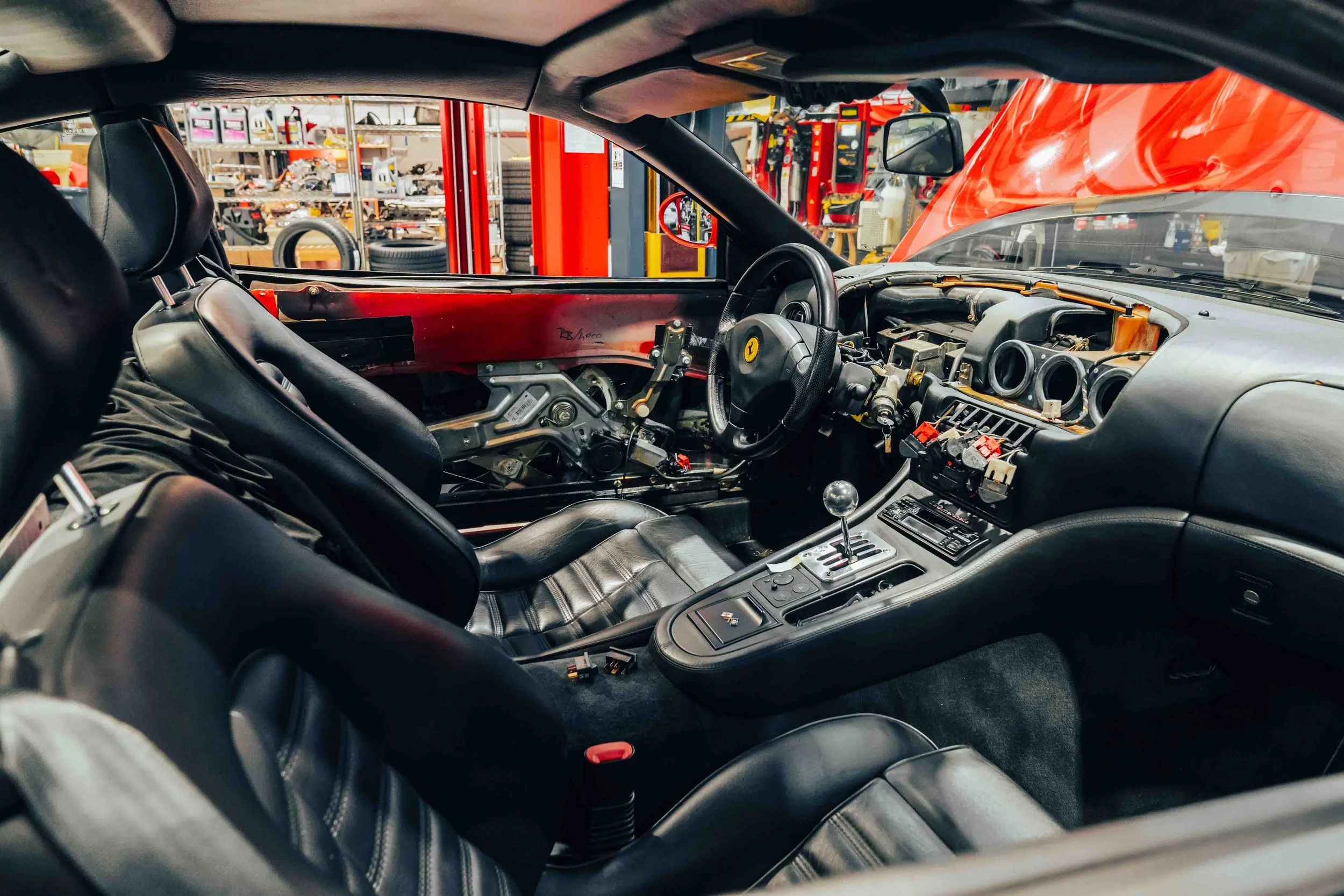Interior of a Ferrari race car with black leather seats, dashboard, and steering wheel, in a garage or workshop.