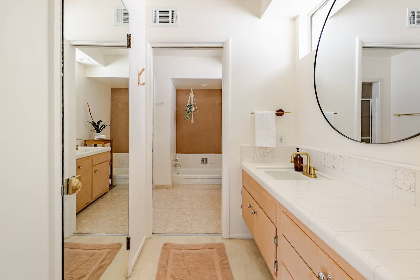 Bright bathroom with white walls, large mirror, and wooden cabinetry with brass fixtures. A sink, a towel hanging on a bar, and a window are visible.