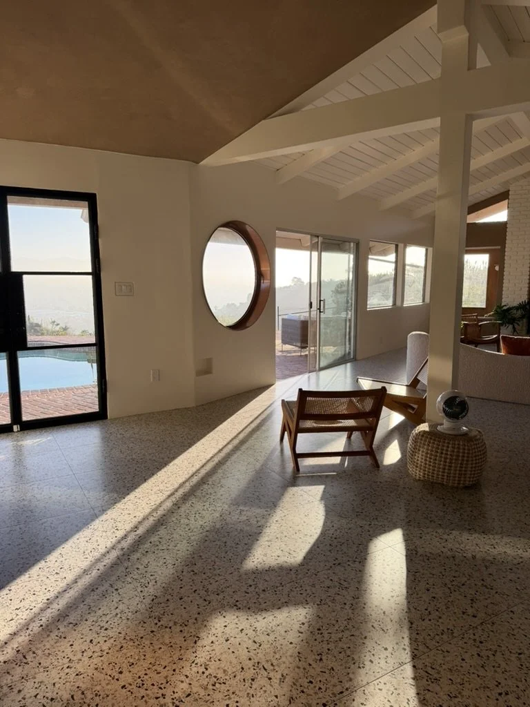 Bright living room with sunlight casting shadows, sliding glass door to outdoor pool, round mirror on white wall, chairs, and windows with scenic view.