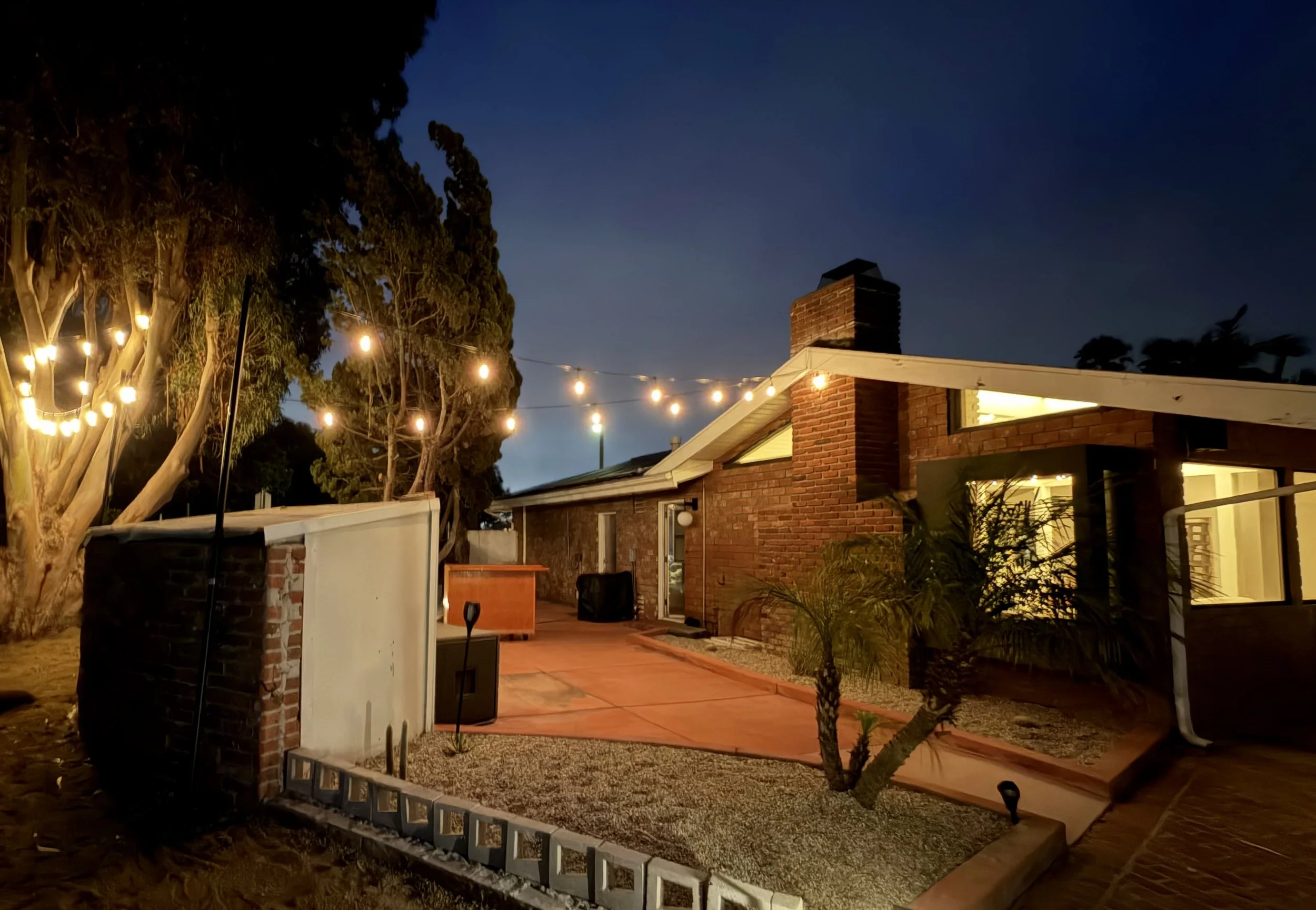Nighttime backyard scene with string lights hanging between trees and house with brick chimney, glass door, and outdoor patio area.