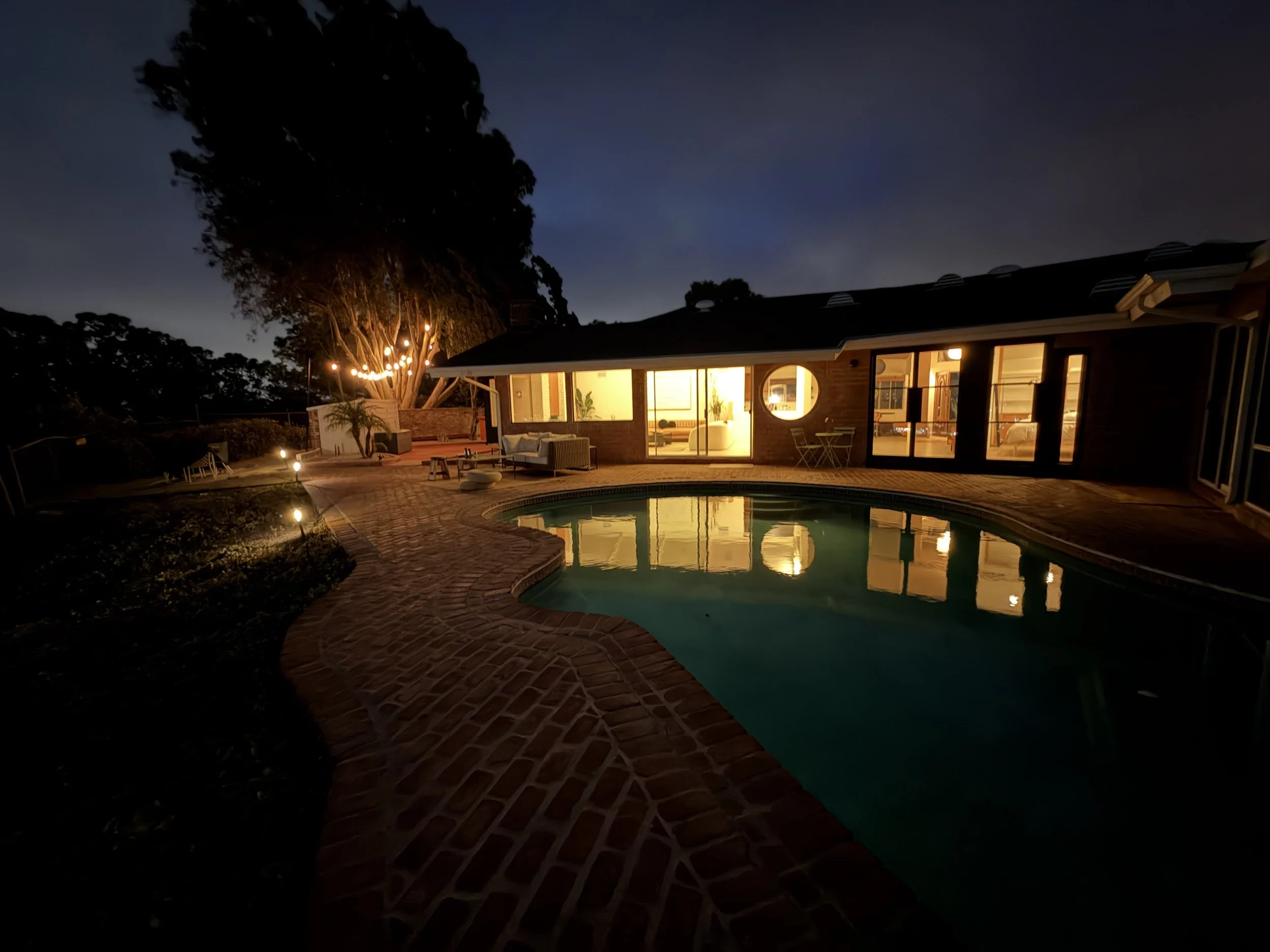 A backyard at night with a swimming pool, outdoor furniture, a large tree with string lights, and a house with illuminated interior.