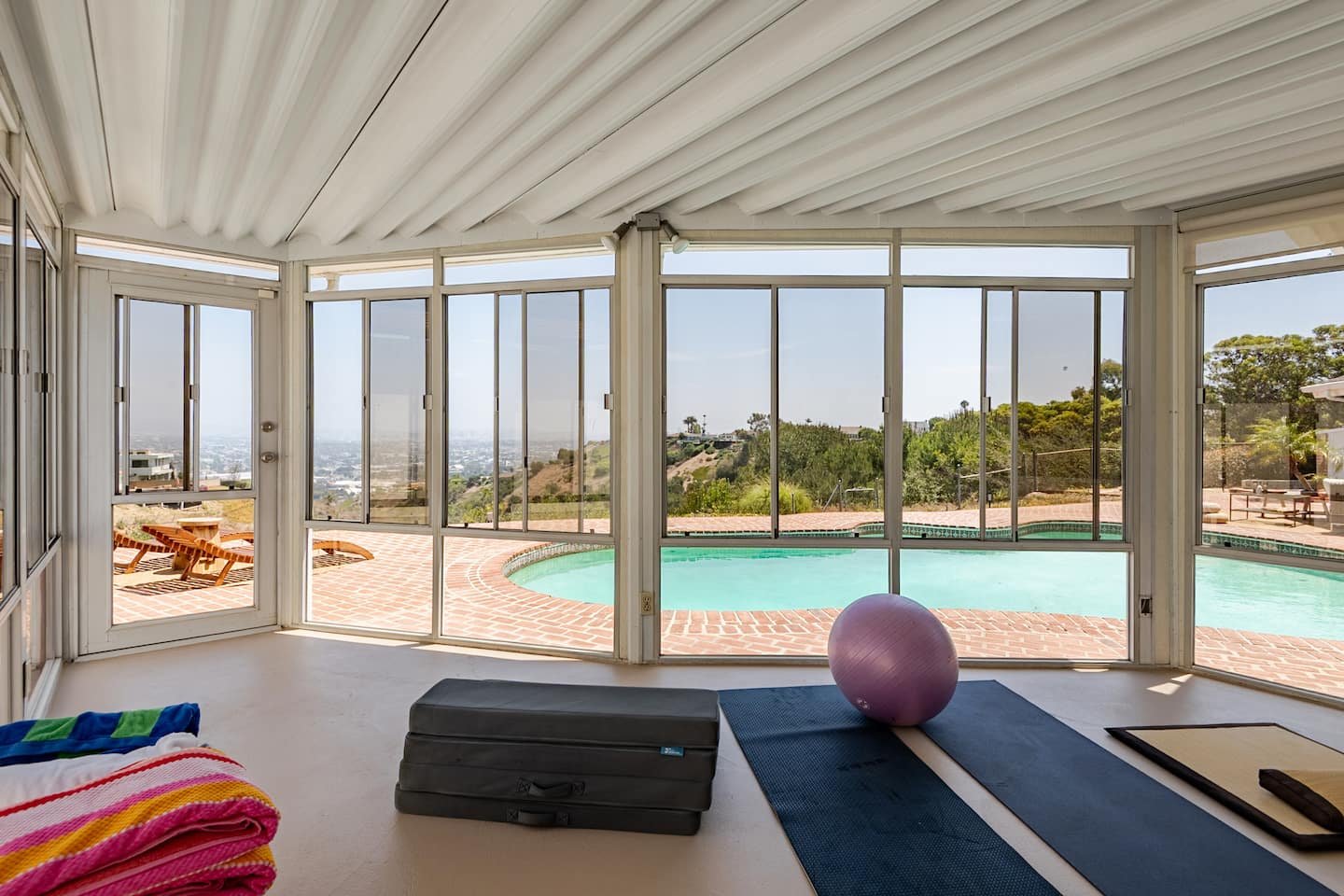 Sunroom with large glass windows and sliding doors, overlooking a private pool with brick surround and hillside view, fitness equipment and towels inside.