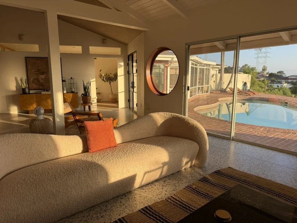 Living room with a cream-colored curved sofa, orange pillow, and large sliding glass doors leading to a pool area outside, with warm sunlight illuminating the space.