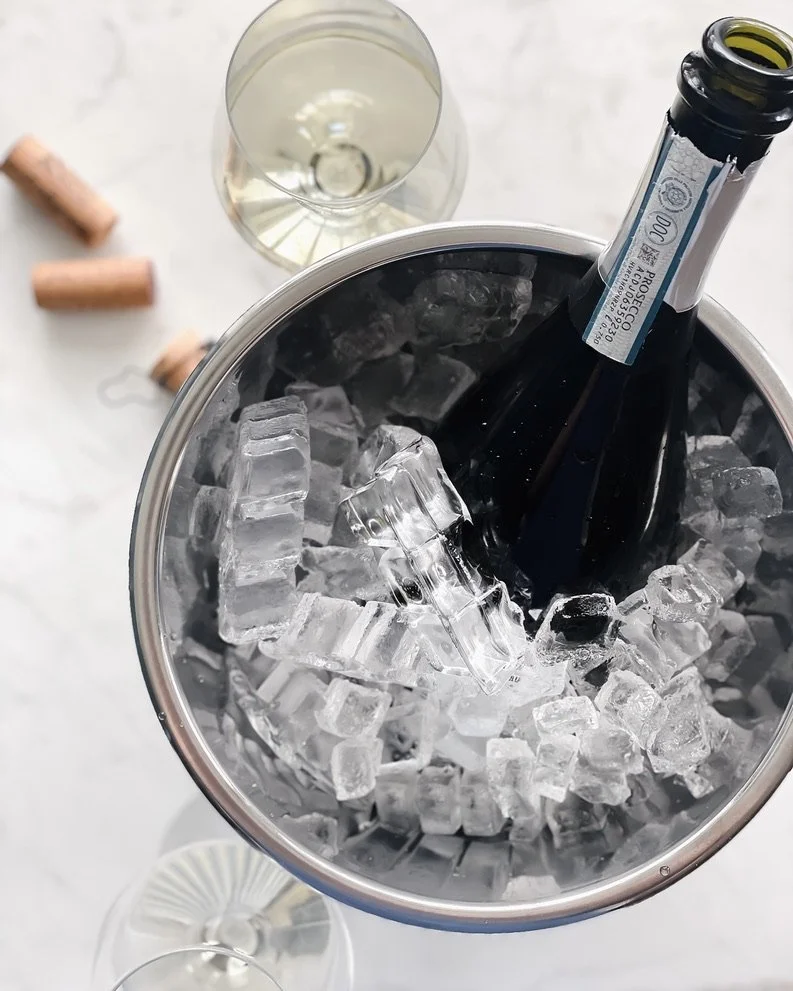 A bottle of champagne chilling in an ice bucket, with a glass of champagne and some corks on a white surface.