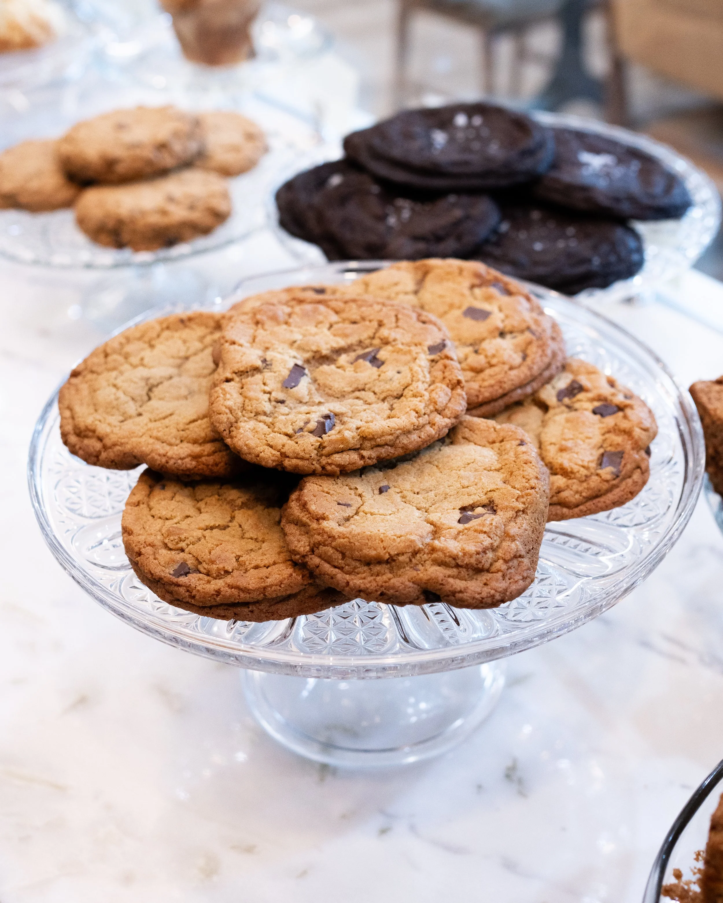Plate of chocolate chip cookies on a glass cake stand with cookies in the background.